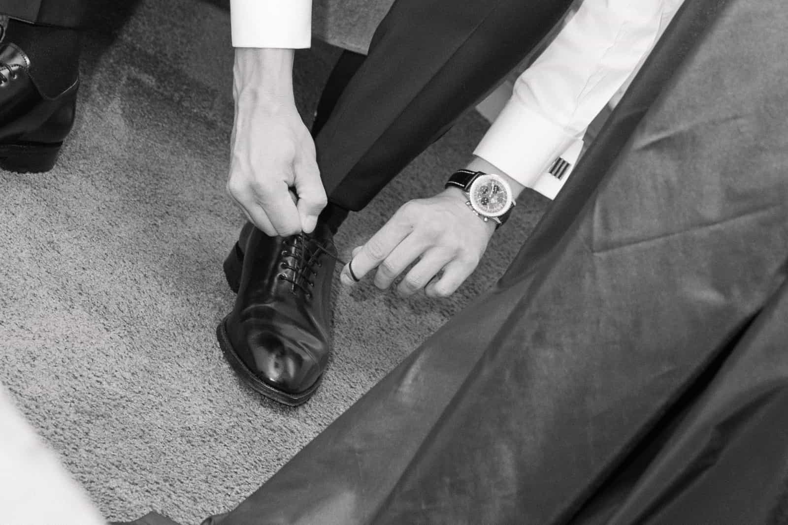 Bräutigam bindet schwarze Oxford-Schuhe mit Luxusuhr sichtbar, Schwarzweiß Getting Ready