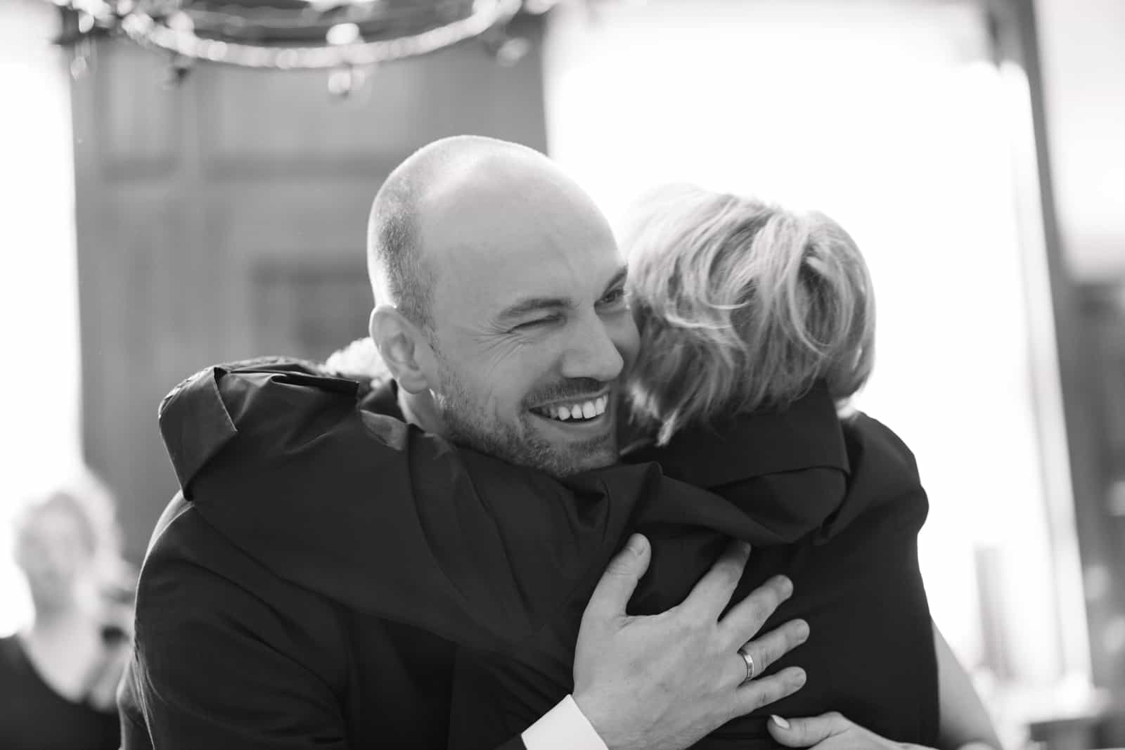 Bräutigam umarmt blonde Gästin mit echtem Lachen, Schwarzweiß