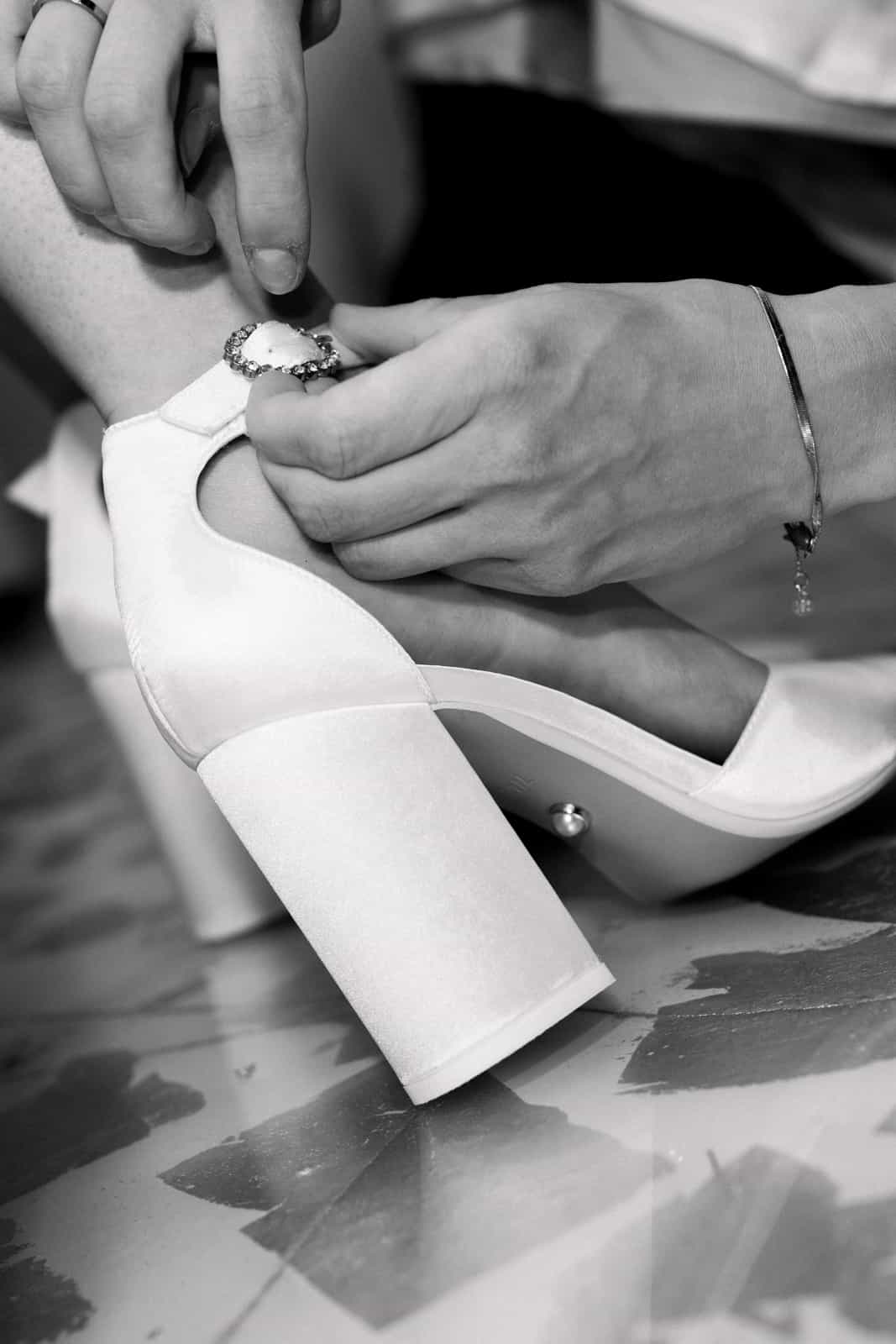 Braut zieht weiße Hochzeitsschuhe an mit Verlobungsring und Armband sichtbar, Schwarzweiß