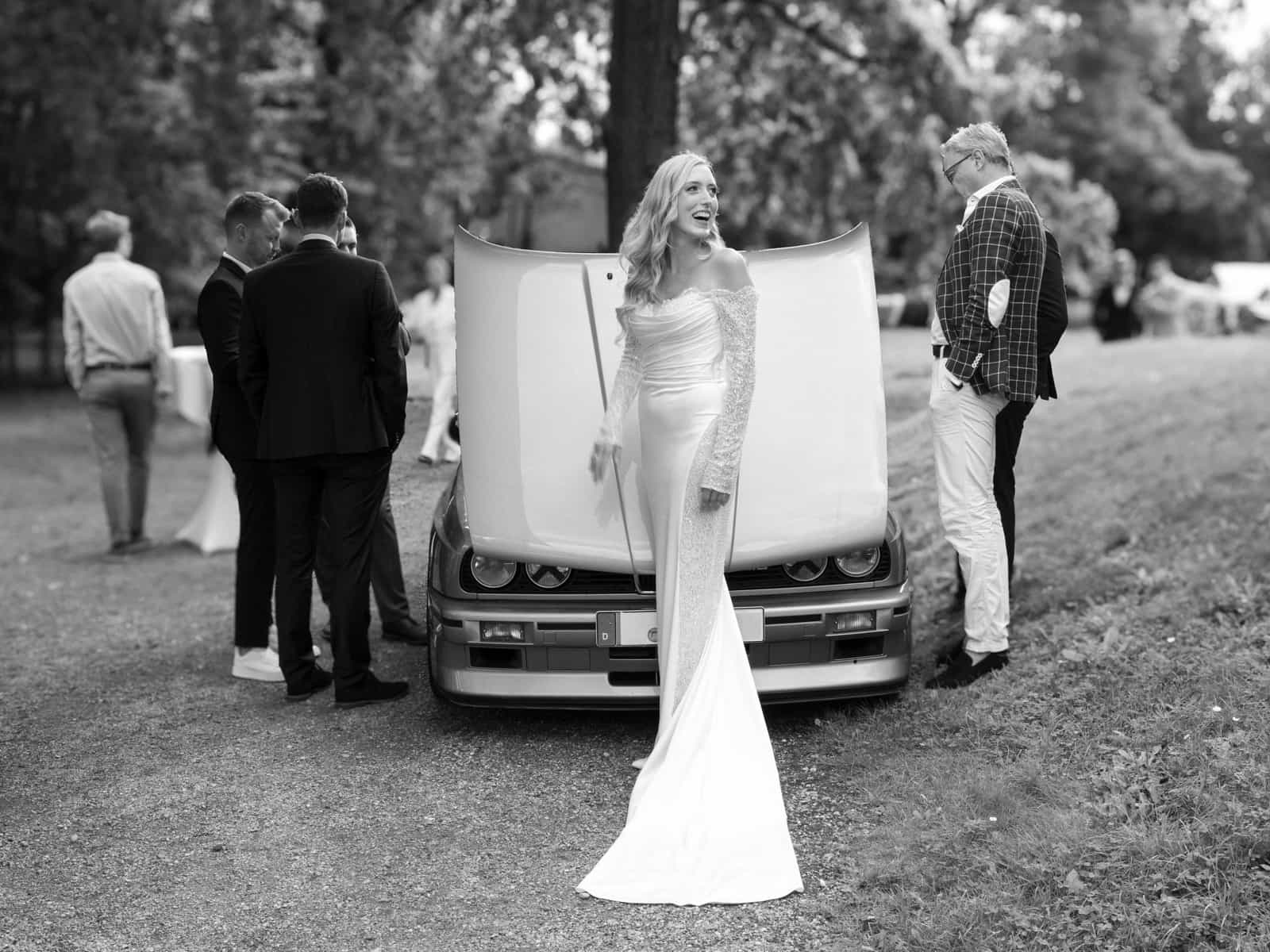 Braut in weißem Brautkleid mit Spitzenärmeln steht vor Vintage-Auto bei Hochzeit Schloss Kartzow
