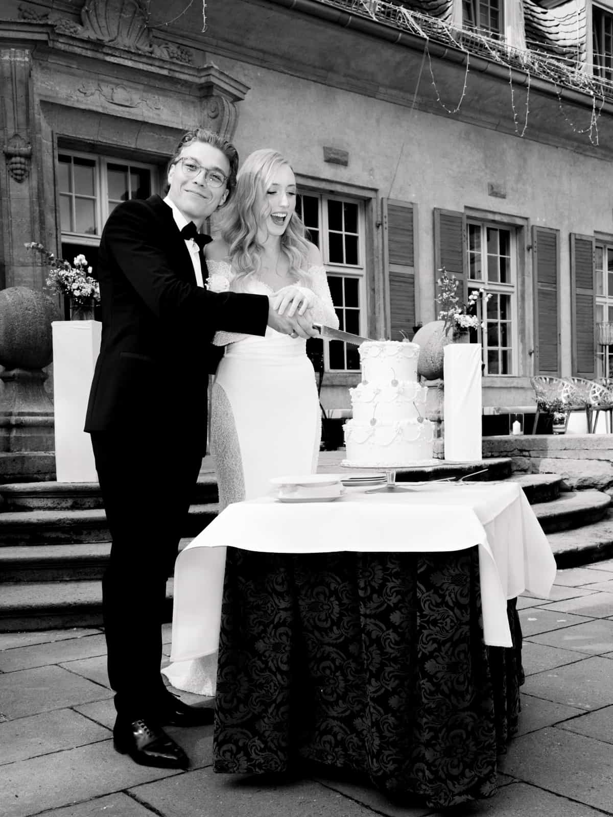 Brautpaar schneidet gemeinsam weiße Hochzeitstorte vor Schloss Kartzow, schwarz-weiß Fotografie