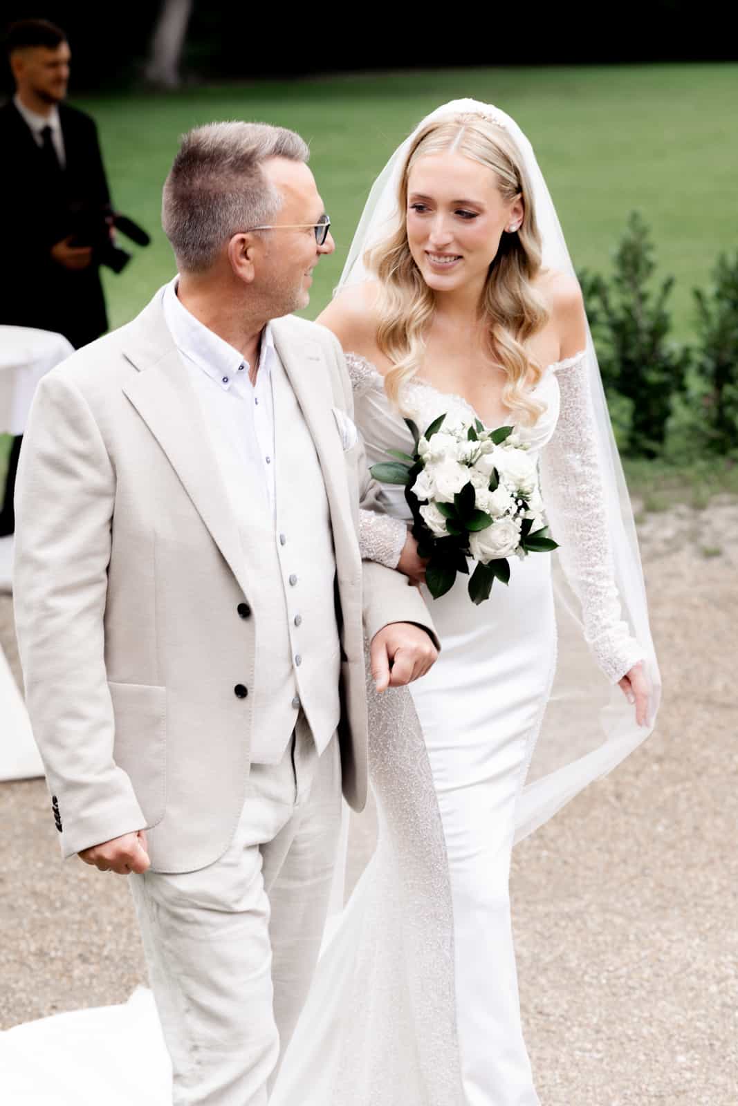 Brautvater in beigem Anzug führt Braut im Spitzenkleid mit Rosenstrauß zum Altar