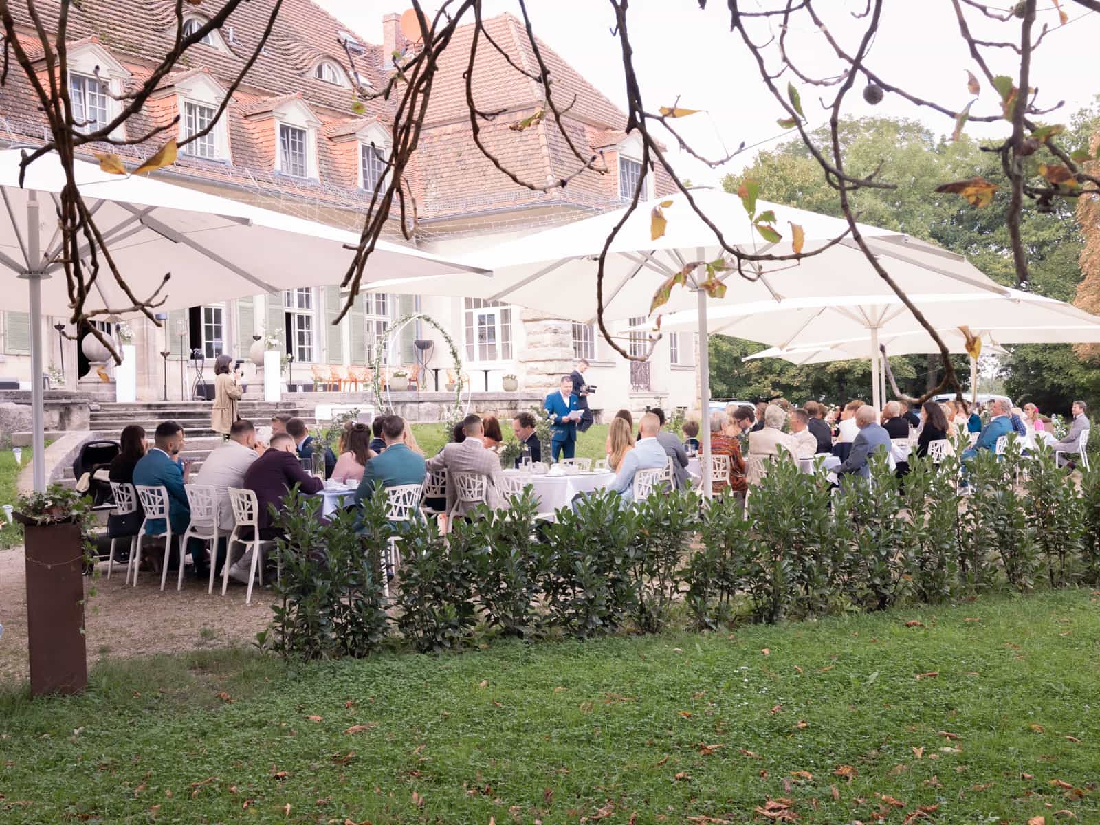 Hochzeitsgäste bei freier Trauung im Garten vor Schloss Kartzow mit weißen Sonnenschirmen