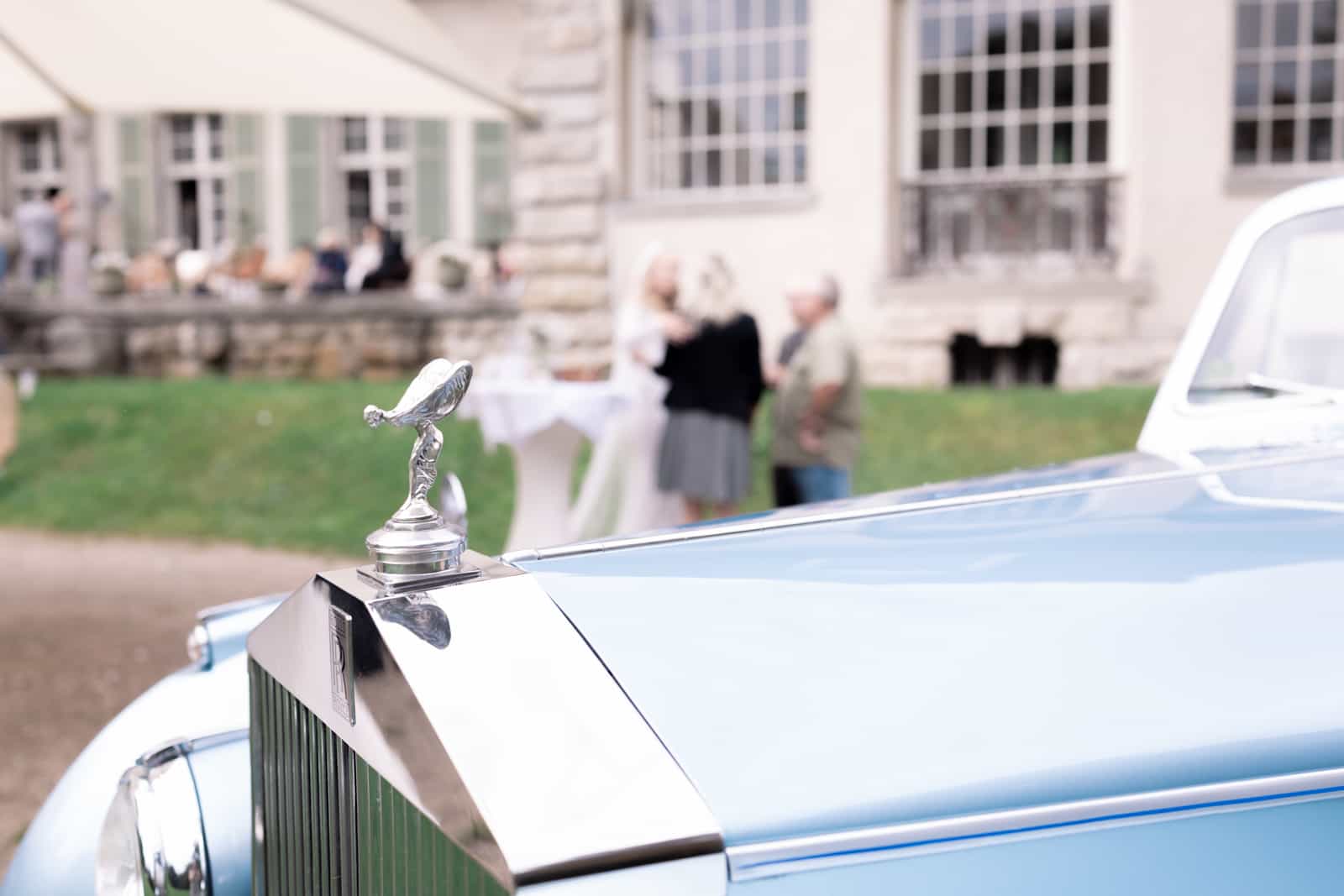 Rolls-Royce Kühlerfigur mit Brautpaar unscharf vor Schloss im Hintergrund