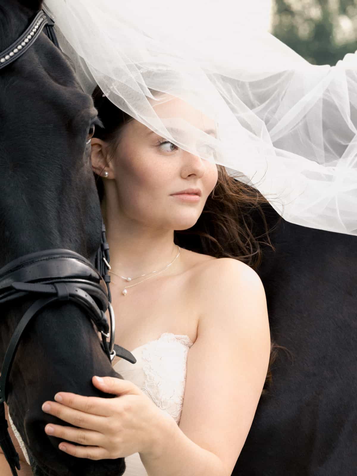 Bride with fluttering veil leaning dreamily against black horse