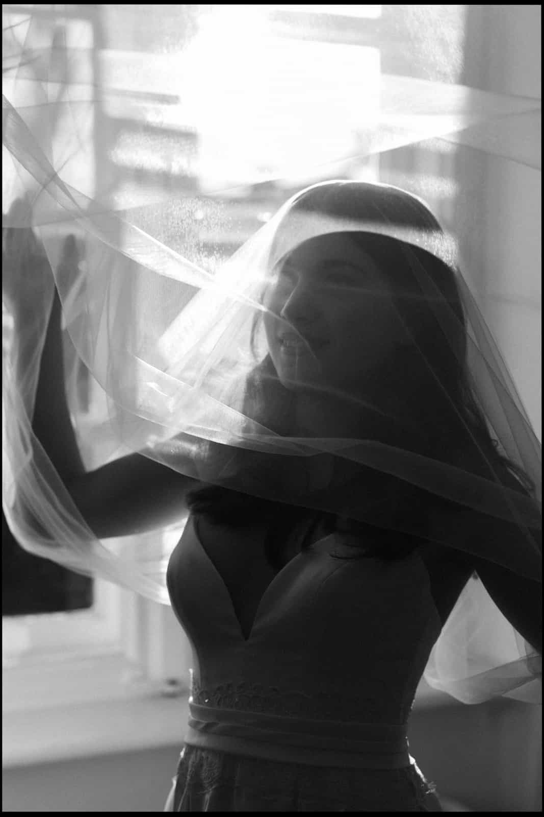 Bride with dramatically swirling veil by window, black and white artistic