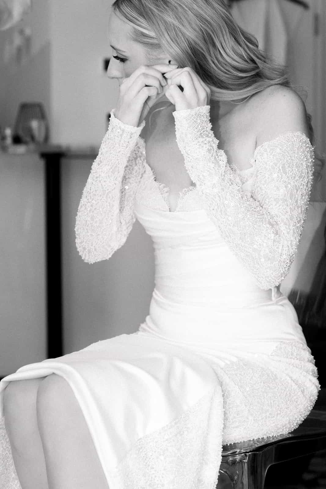 Bride putting on earrings during getting ready in wedding dress with lace sleeves, black and white