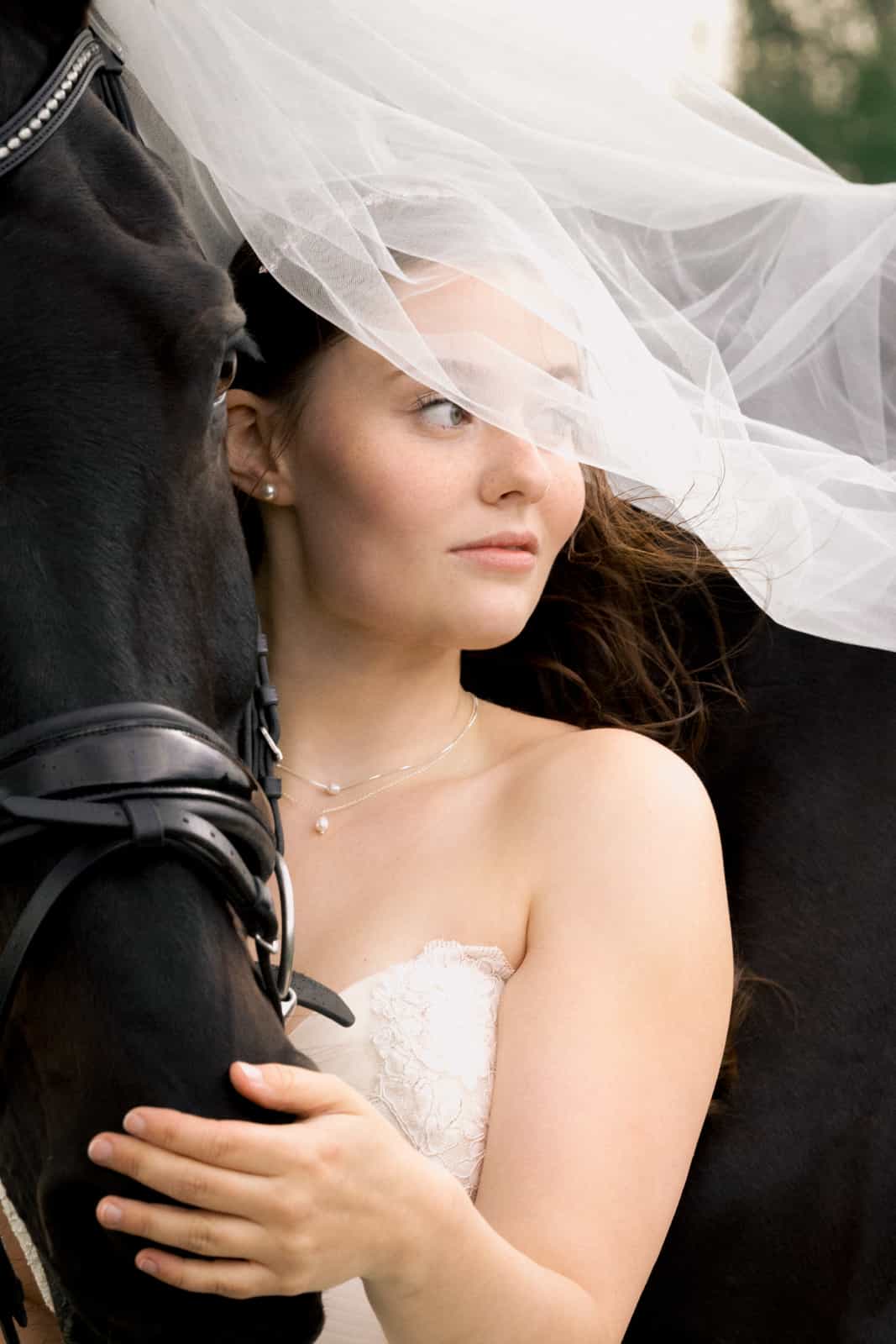 Bride with veil embracing black horse with bridle, pearl necklace visible