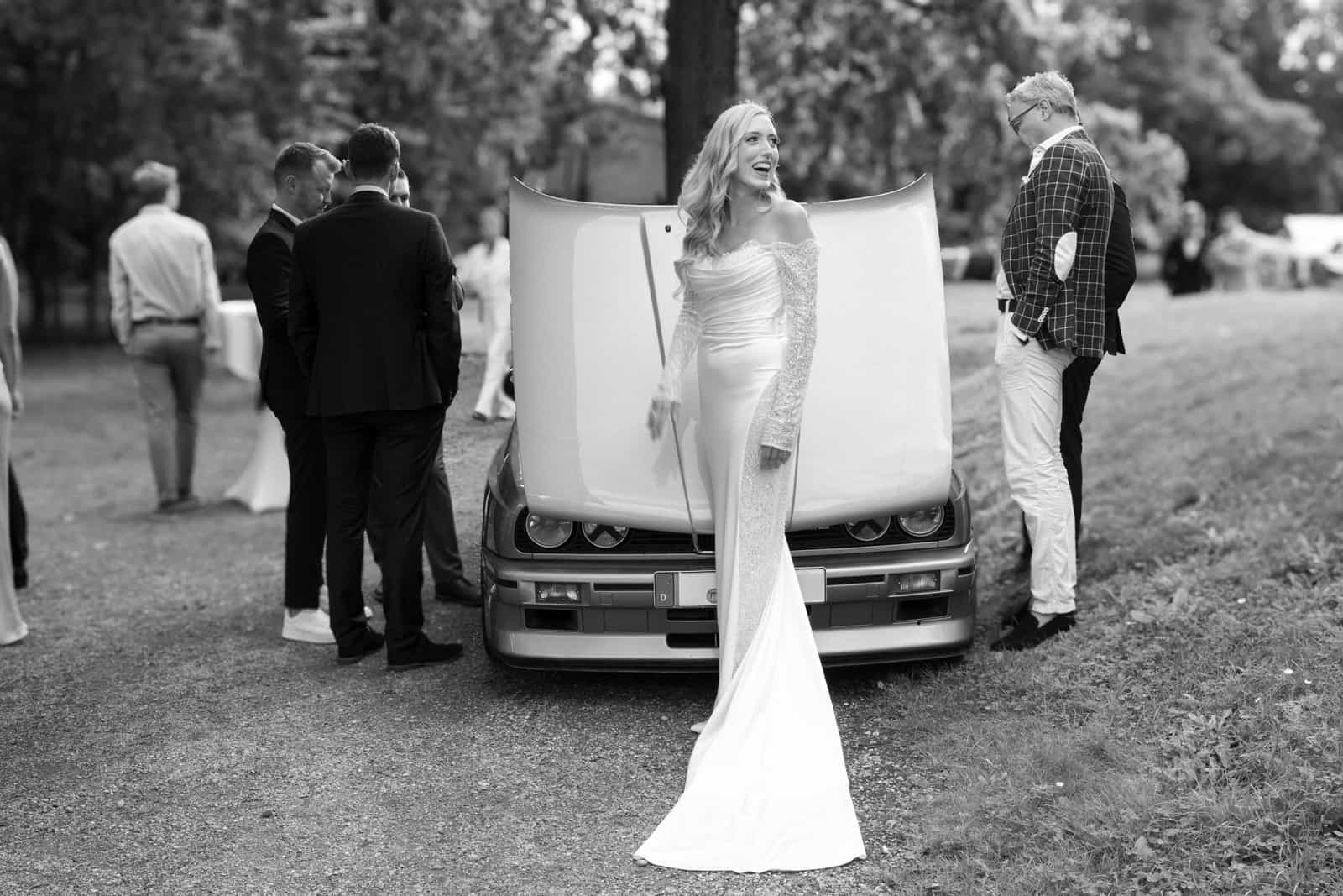 Bride in fitted wedding gown laughing beside vintage BMW on estate grounds, black and white