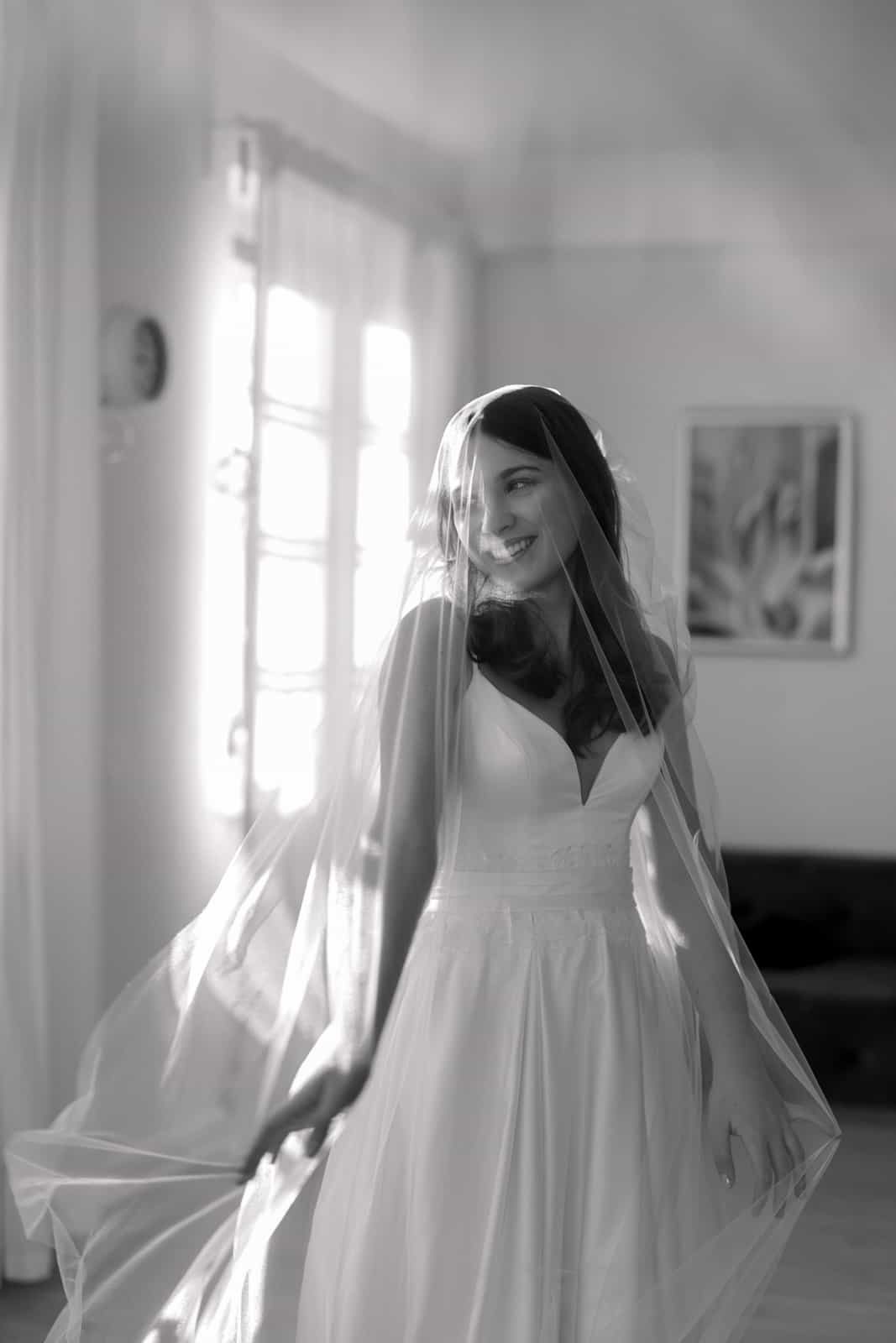 Laughing bride twirling with flowing veil by window, black and white