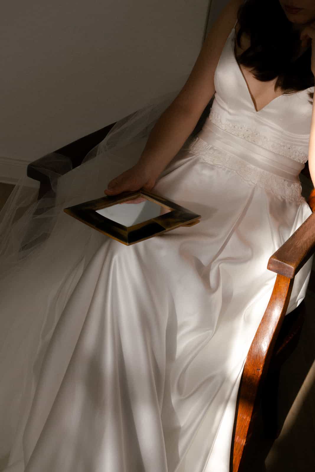 Bride in satin wedding dress with vintage mirror on antique chair