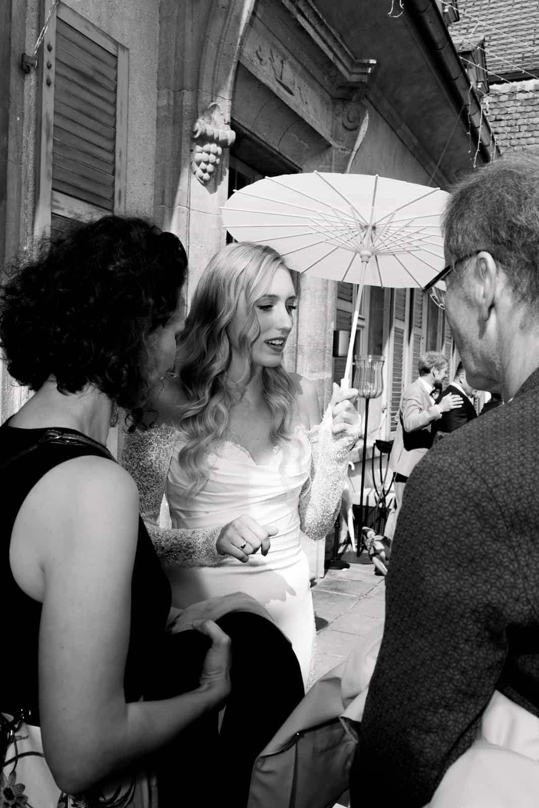 Bride in white wedding dress with parasol talking to wedding guests in front of church in black and white