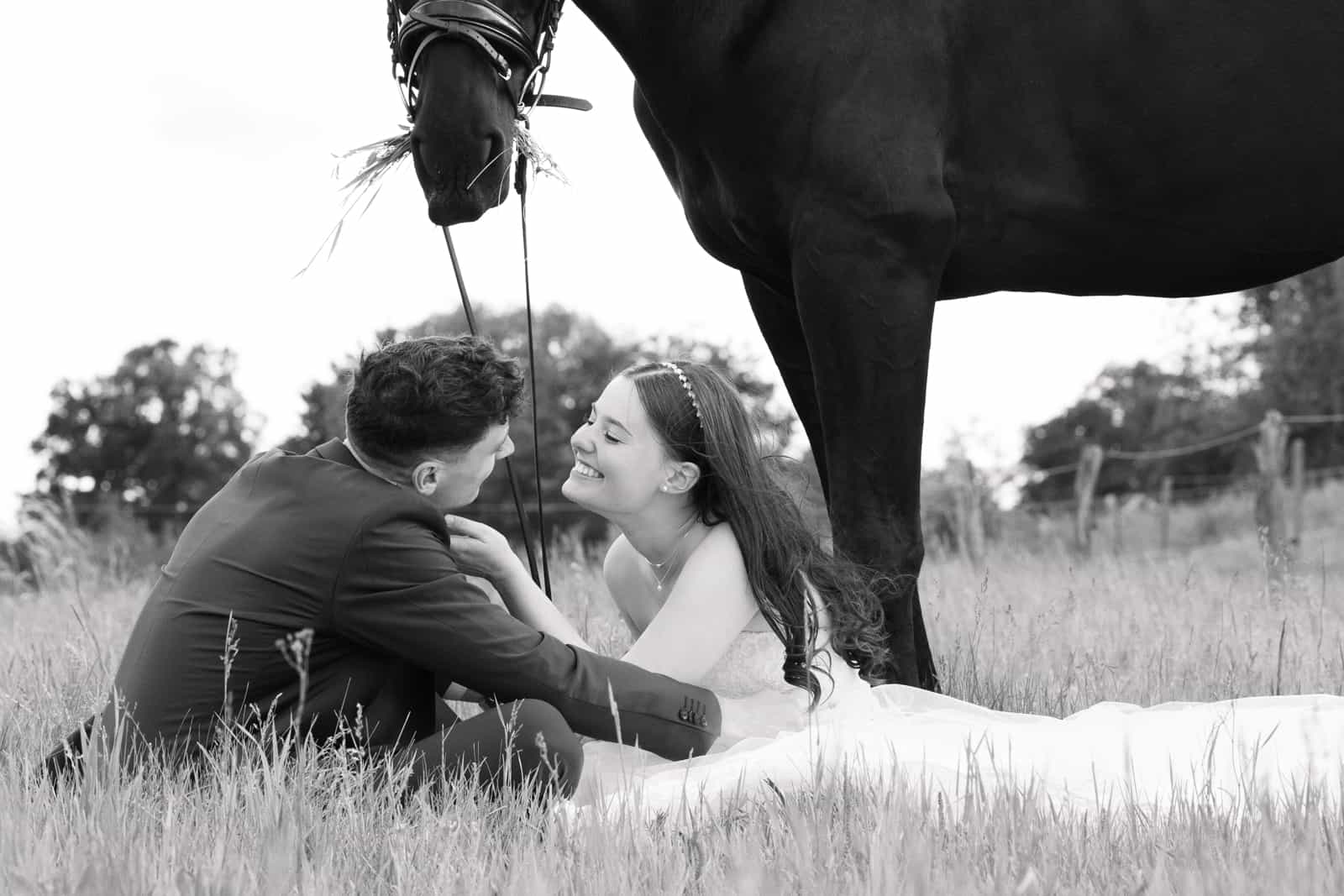 Couple sitting in meadow with black horse standing above, black and white