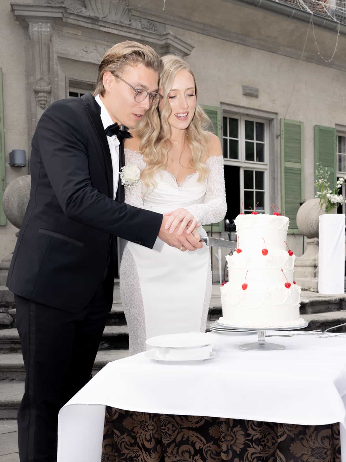 Bride and groom cutting three-tier white wedding cake decorated with red berries in front of Schloss Kartzow