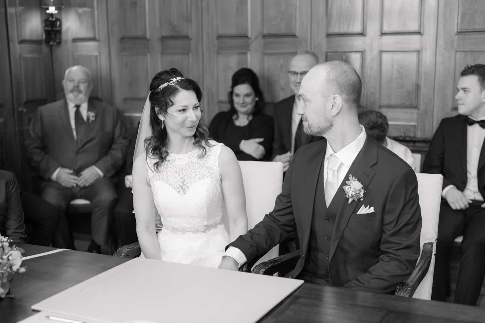 Couple holding hands at civil ceremony, surrounded by guests, black and white