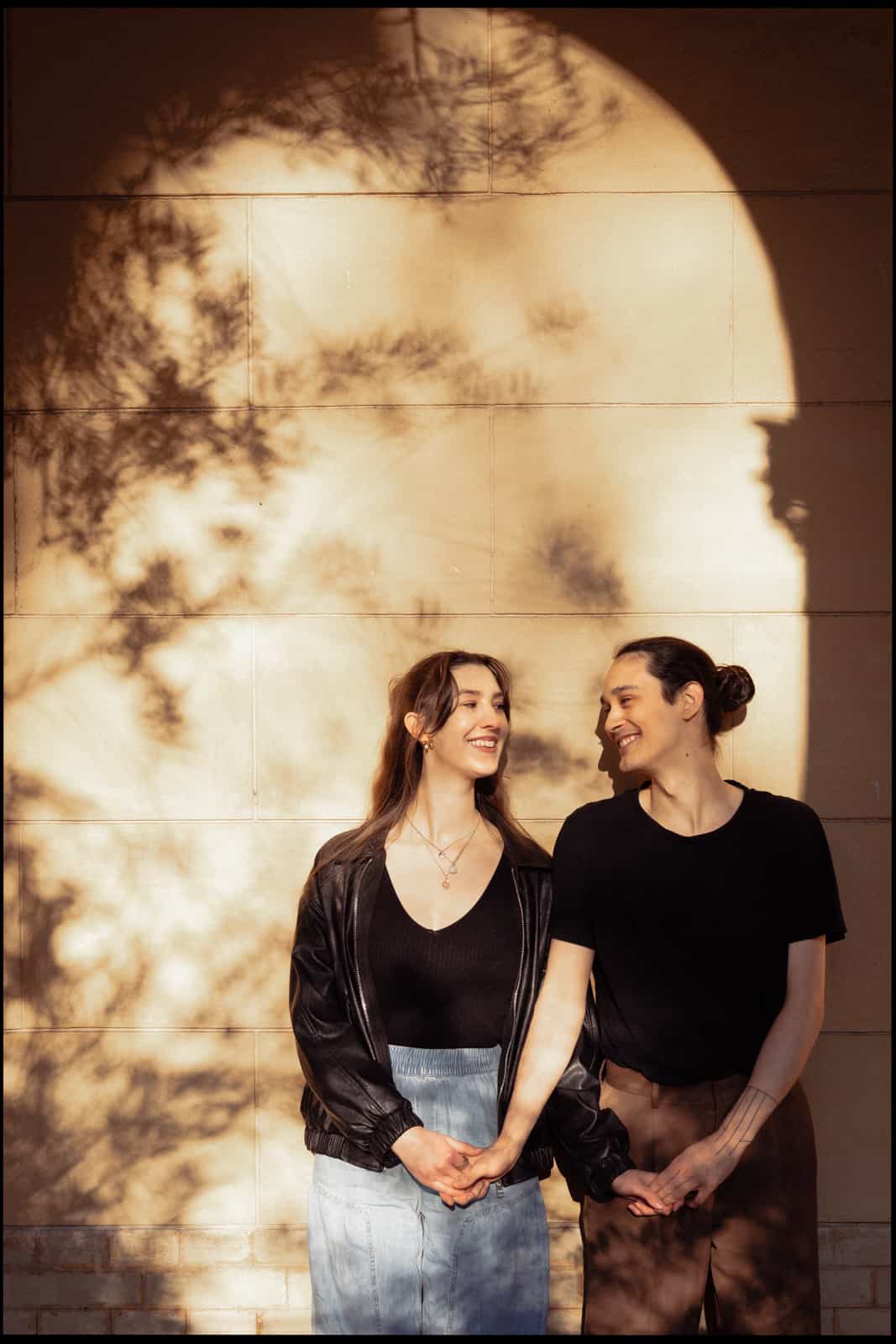 Smiling couple holding hands against a sunlit sandstone wall with foliage shadows