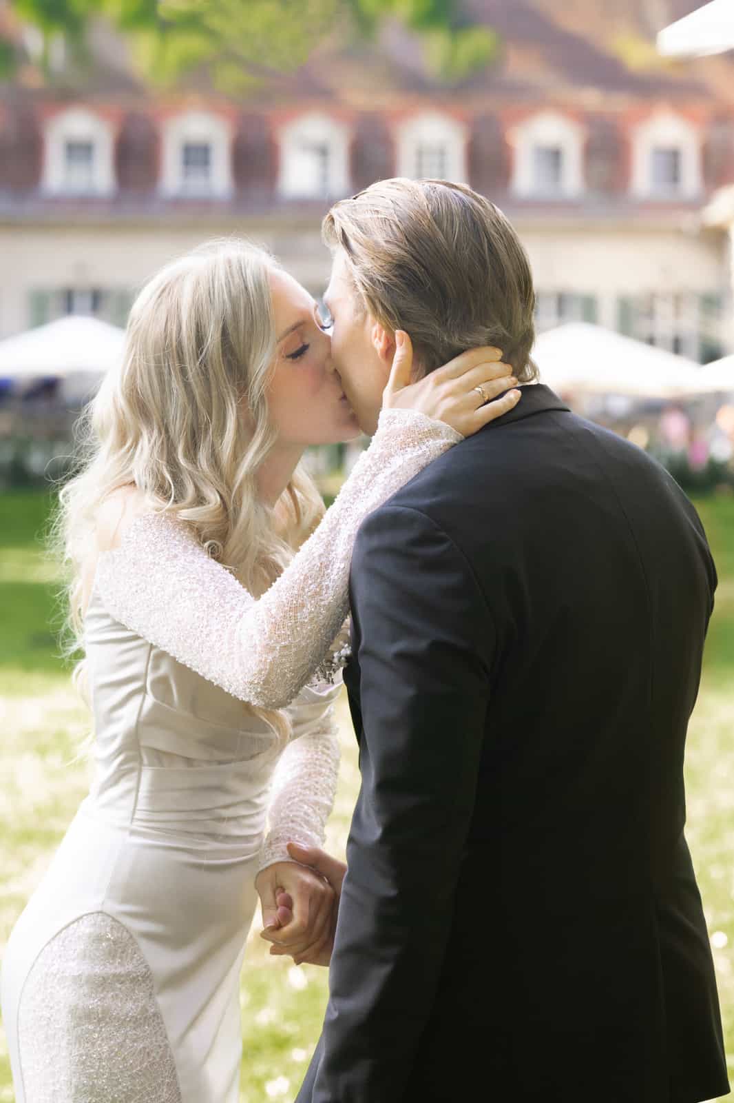 Bride in lace dress kissing groom in garden in front of manor house