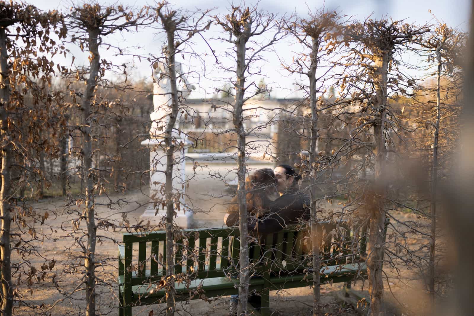 Couple on green park bench seen through bare branches in golden afternoon light