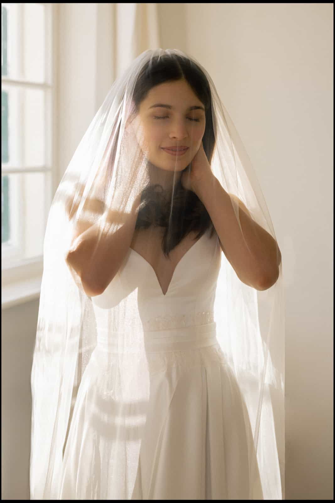 Elegant bride with closed eyes gently touching neck under flowing veil