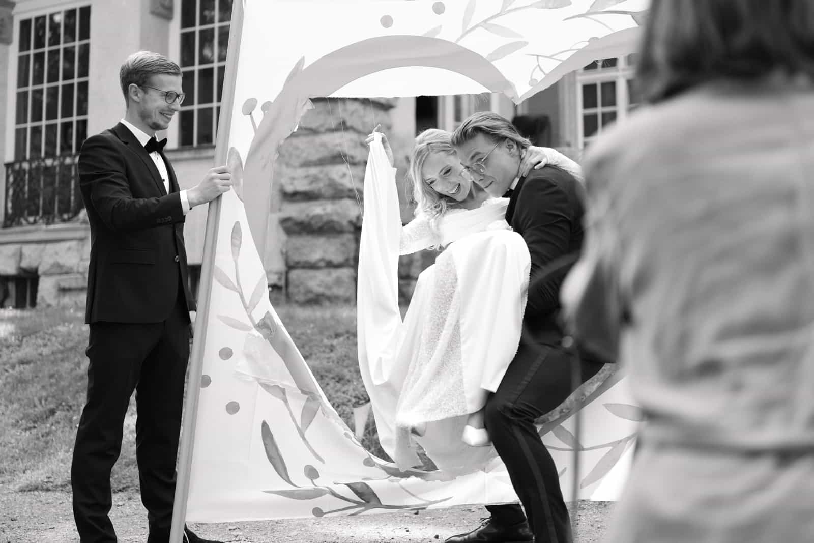 Groom carrying laughing bride through paper heart in German wedding tradition, black and white