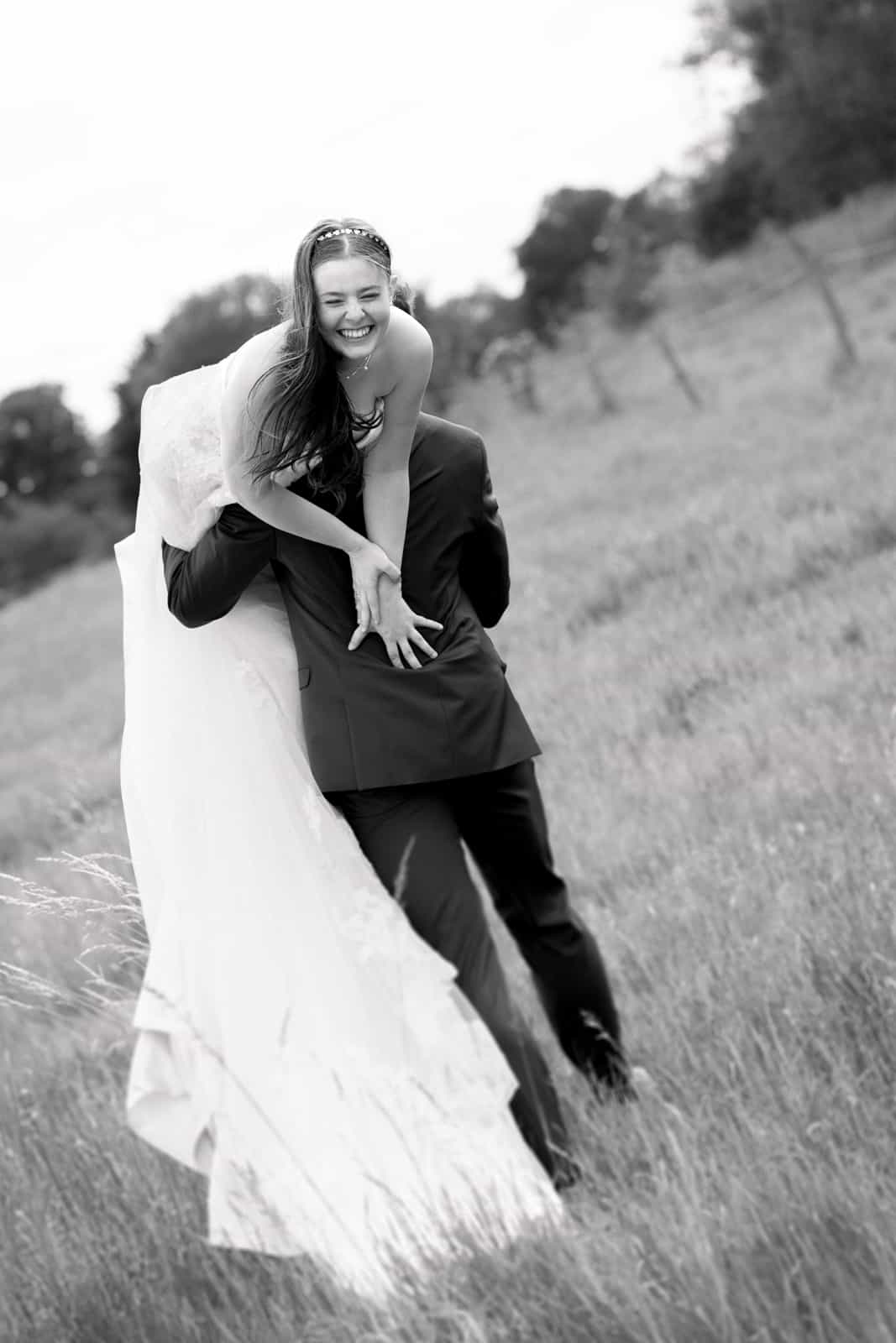 Groom carrying laughing bride piggyback through tall grass, black and white