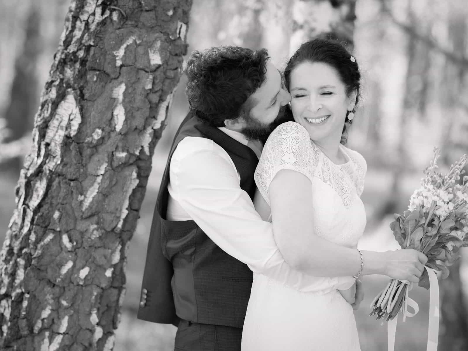 Groom kissing laughing bride on cheek by birch tree, black and white