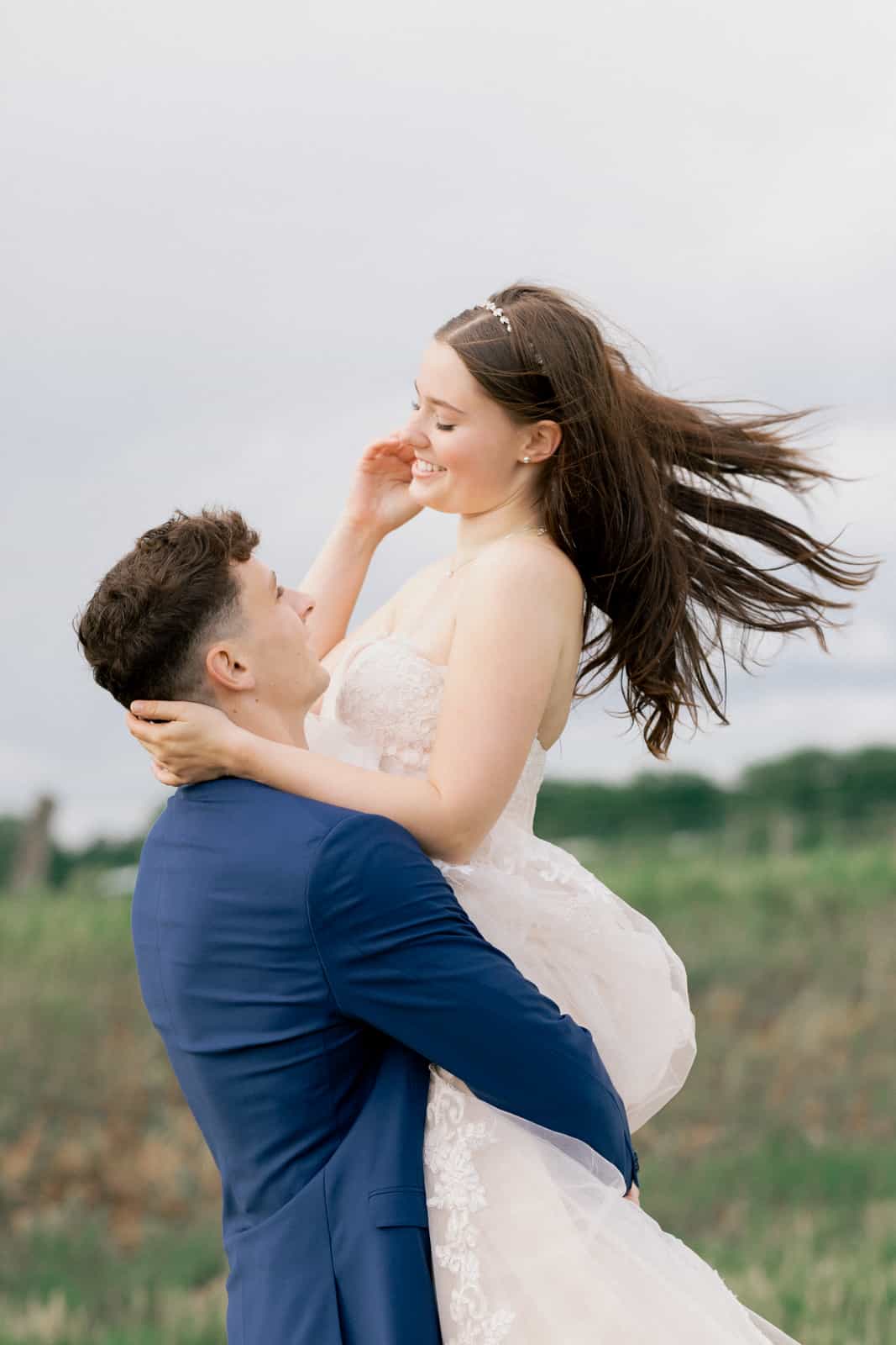 Groom lifting bride high in meadow, windswept hair, intimate eye contact