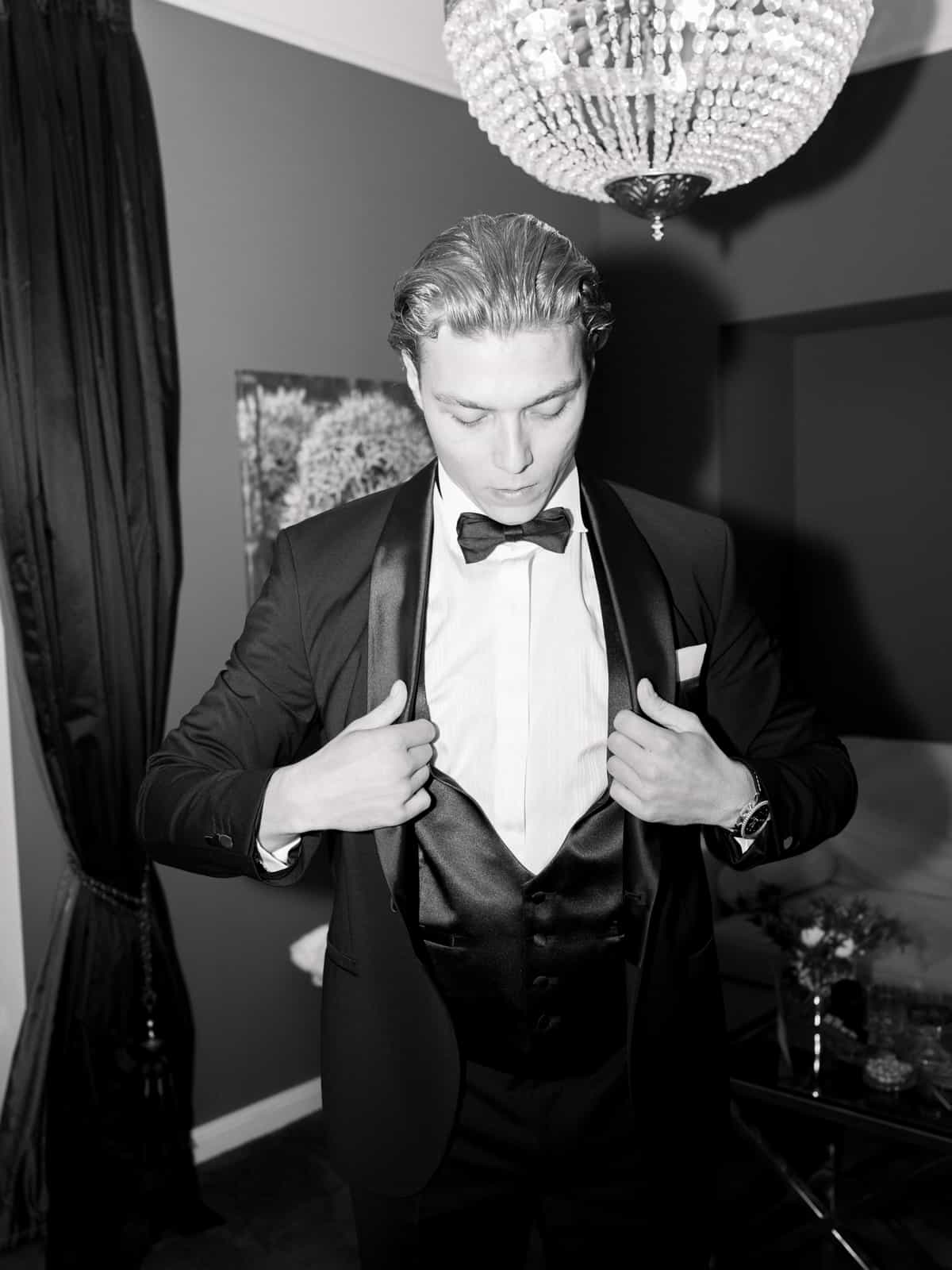 Groom putting on tuxedo jacket under chandelier, getting ready photography Schloss Kartzow