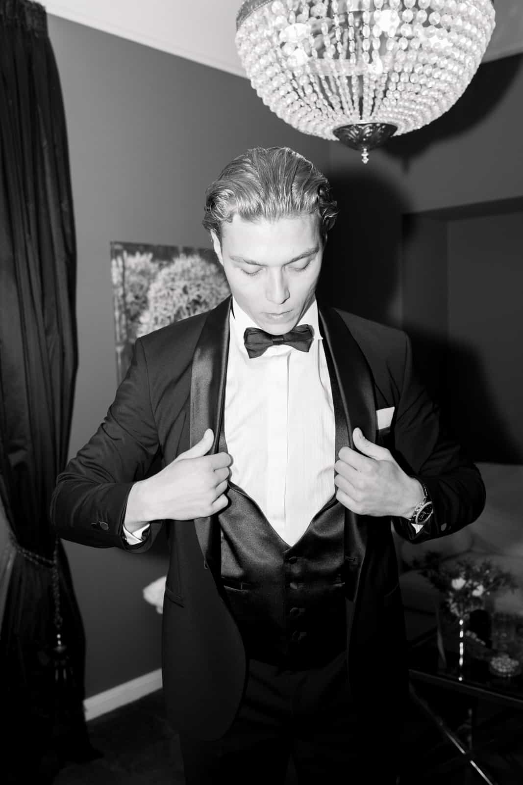 Groom in black tuxedo with bow tie during wedding preparation under chandelier, black and white