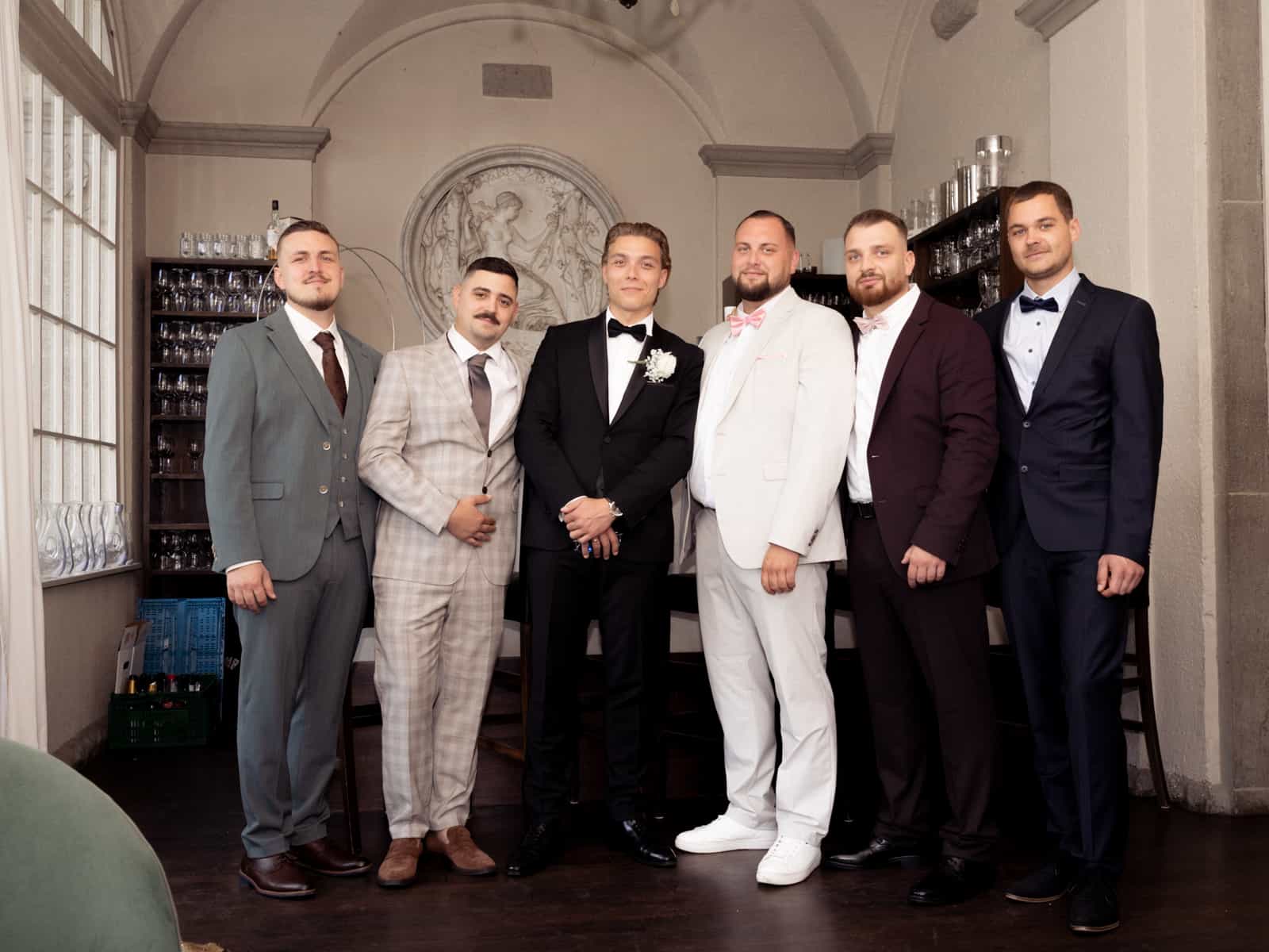 Groom and six groomsmen in suits standing in front of historic wall relief in elegant bar at Schloss Kartzow wedding