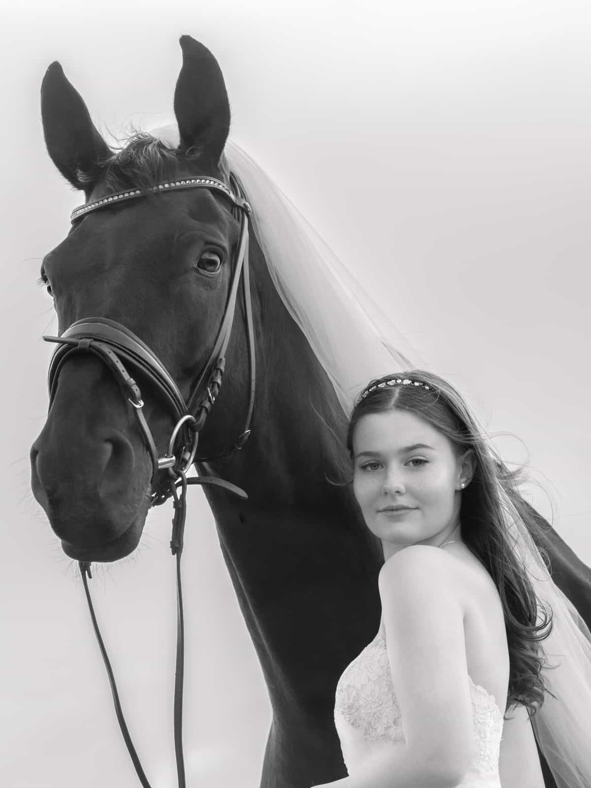 Black and white portrait: Bride with pearl headband next to horse with white veil