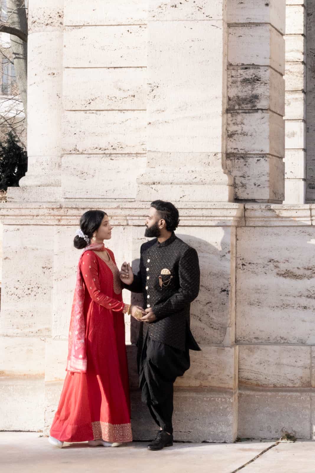 Indian wedding couple in red sari and black sherwani holding hands in front of light stone column