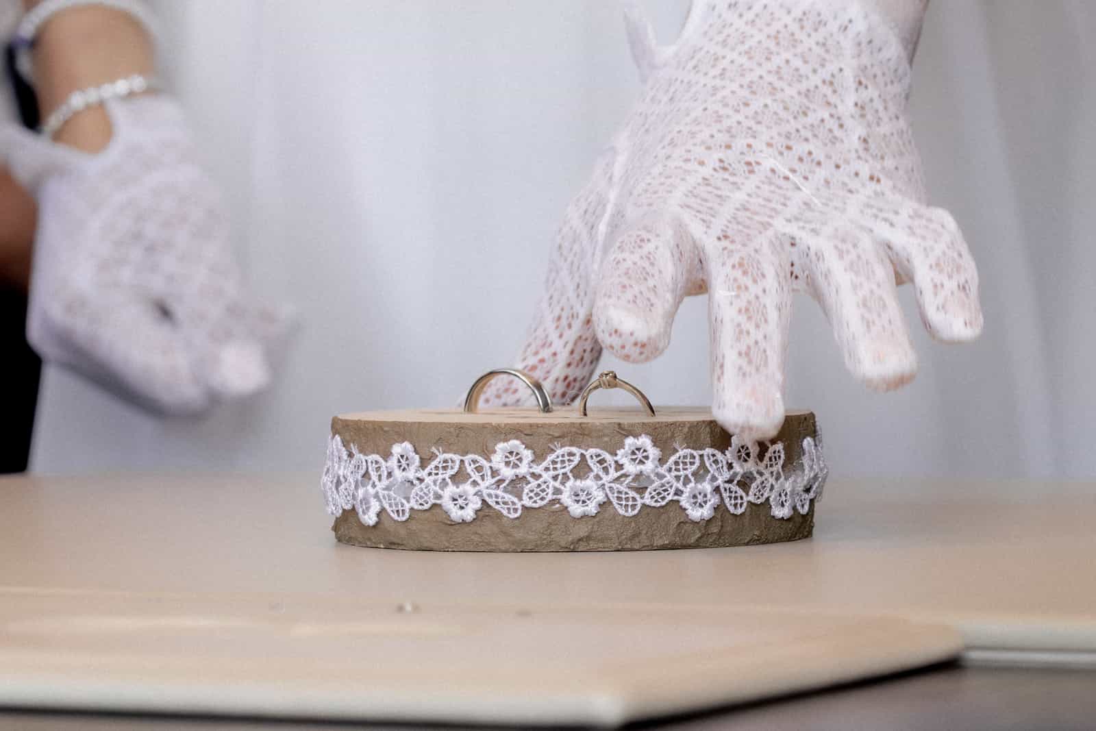 White lace gloves reaching for wedding rings on wooden holder with lace