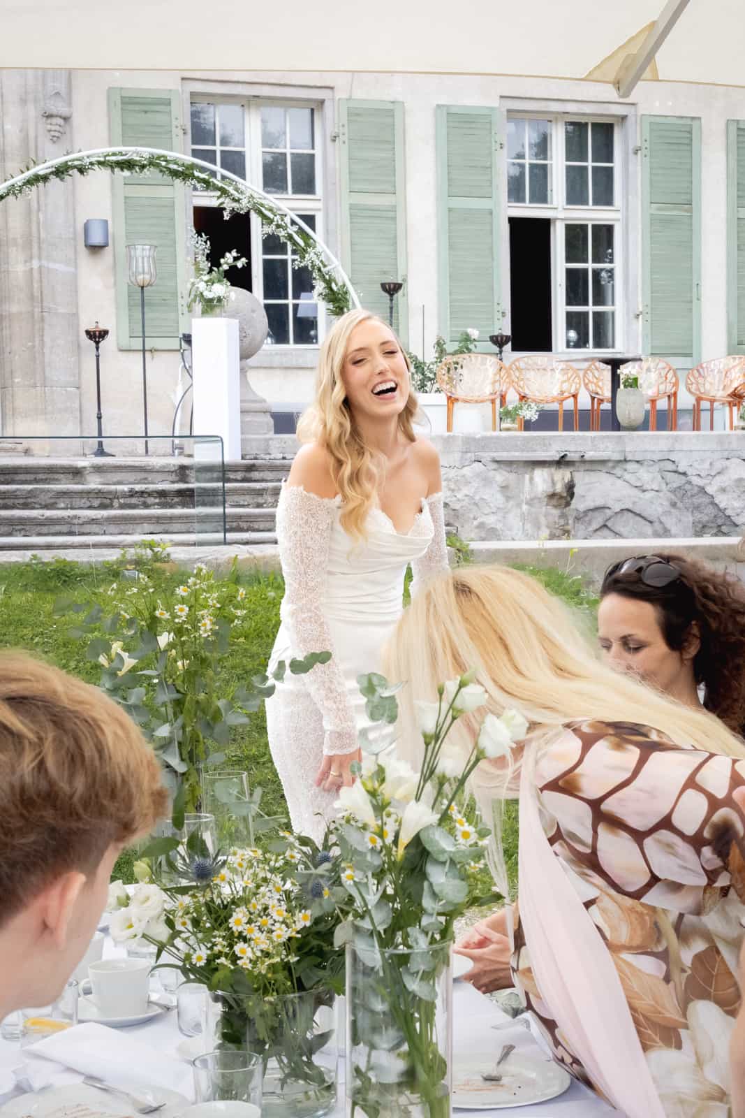 Laughing bride in white lace dress with guests at garden reception in front of manor house