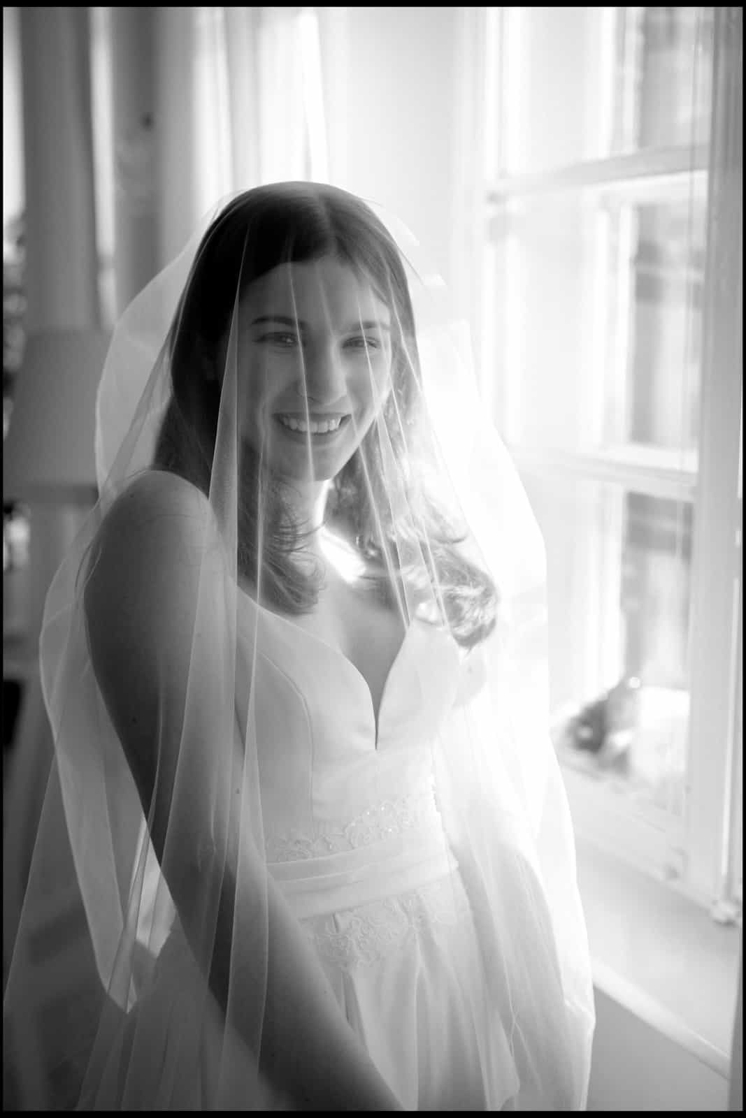Radiantly smiling bride under veil at window, black and white