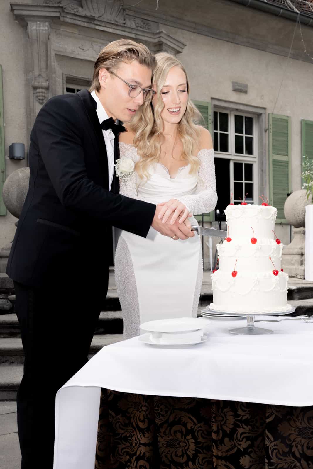 Couple cutting white wedding cake with cherries in front of manor house with green shutters