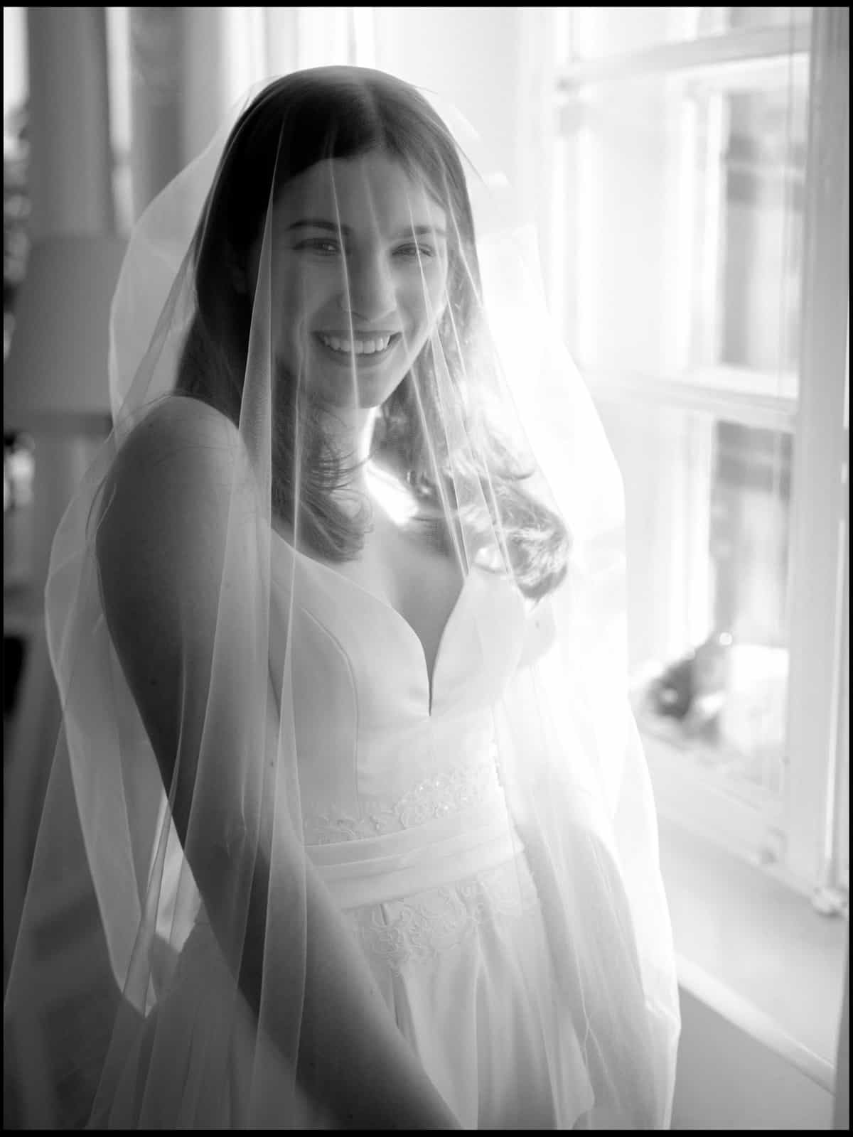 Radiantly smiling bride with veil by window in black and white