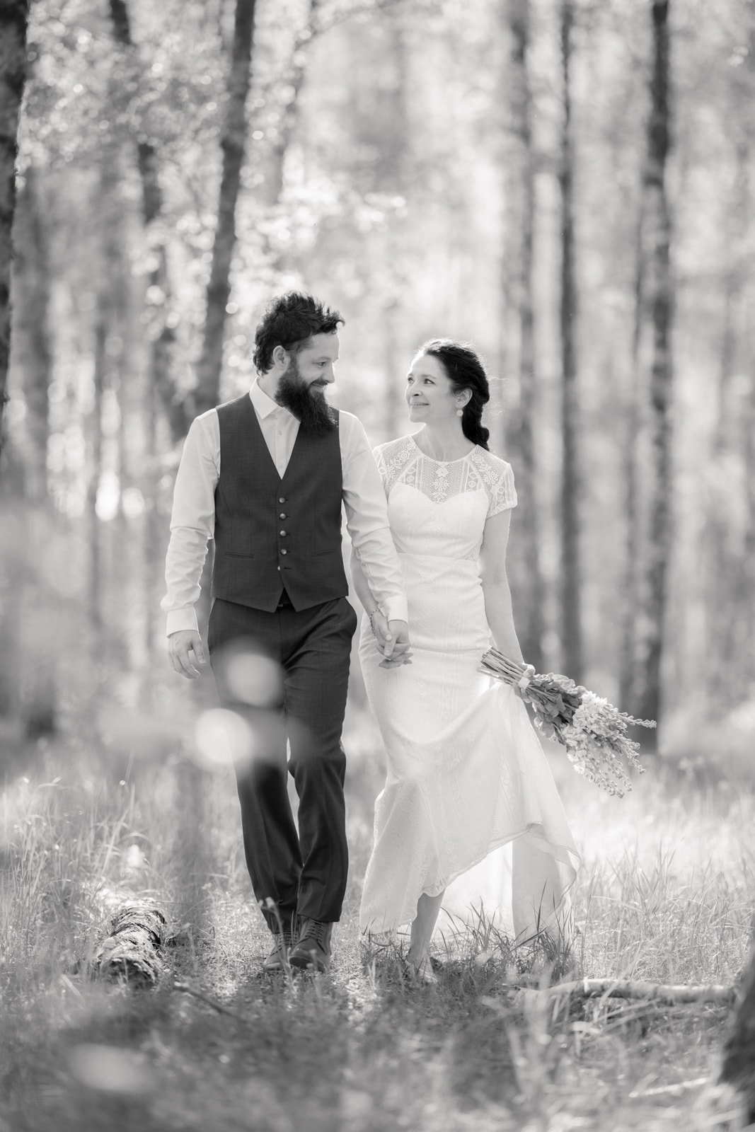 Wedding couple in white dress and vest walking hand in hand through sunny forest