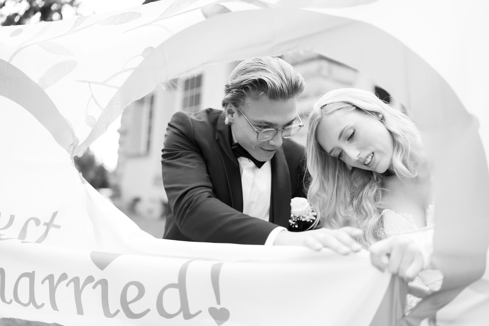 Bride and groom behind a flowing 'just married!' banner, black and white