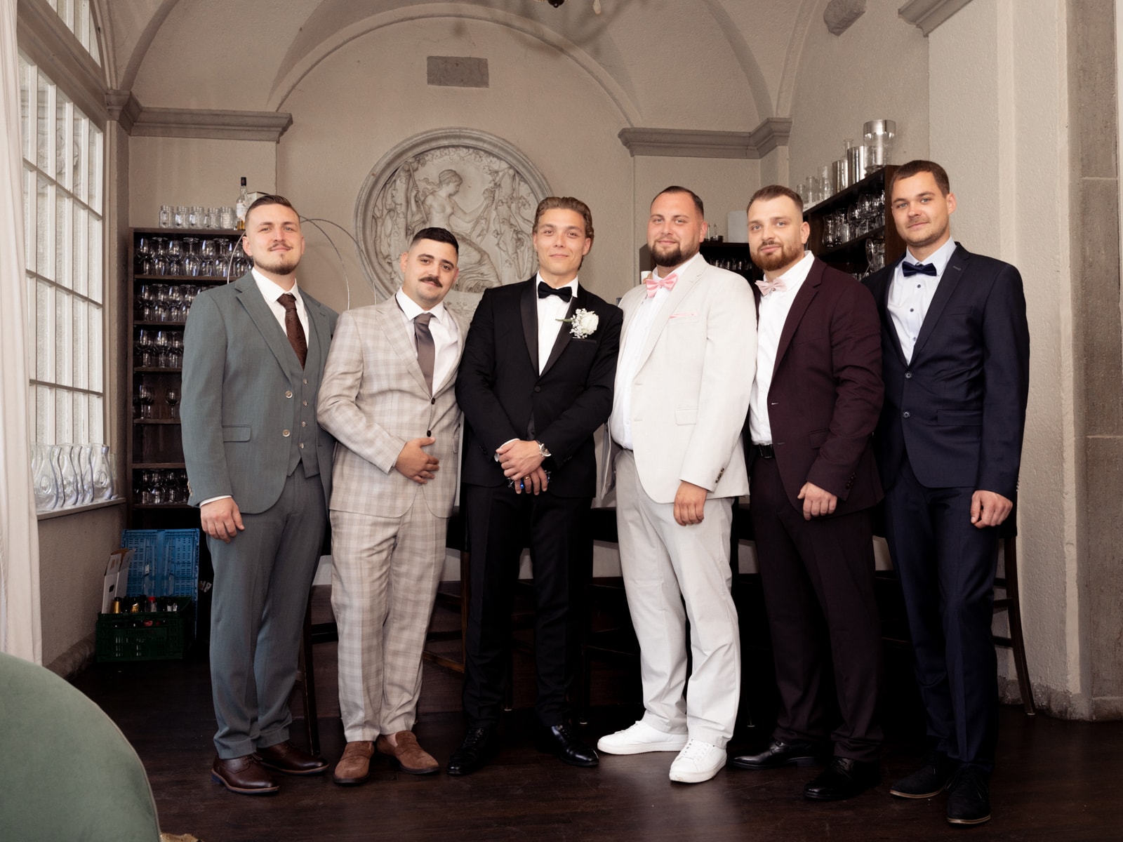 Groom and six groomsmen in suits standing in front of historic wall relief in elegant bar at Schloss Kartzow wedding