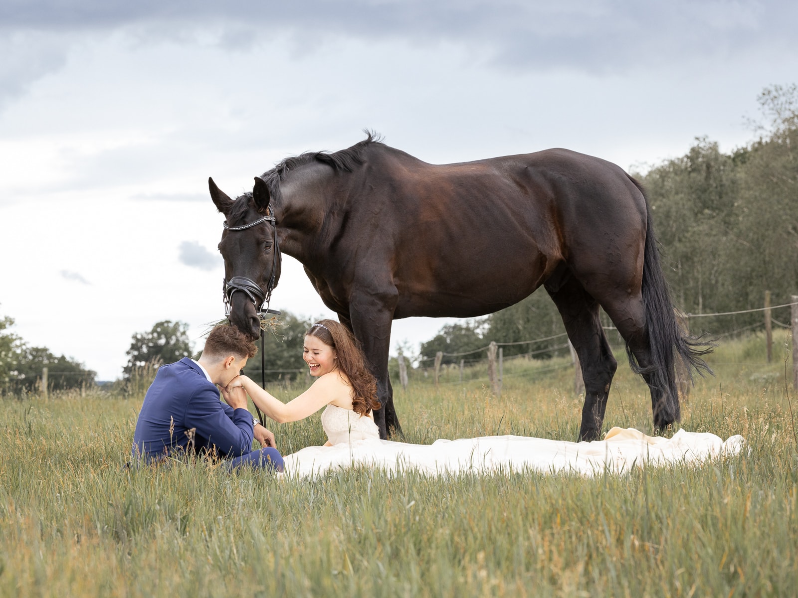 Laughing couple sitting on meadow, groom kissing bride's hand while horse watches