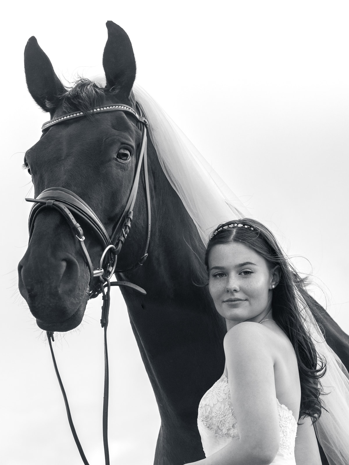 Black and white portrait: Bride with pearl headband next to horse with white veil