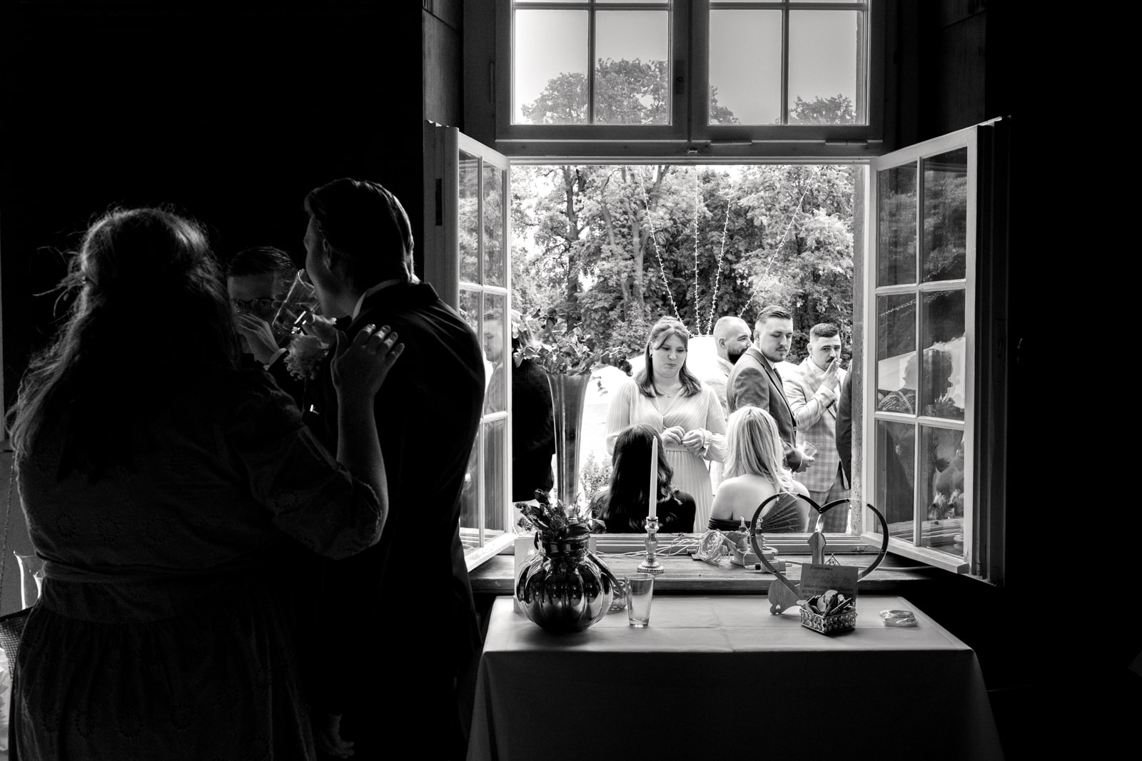 Bride and groom embracing by window while wedding guests celebrate in garden