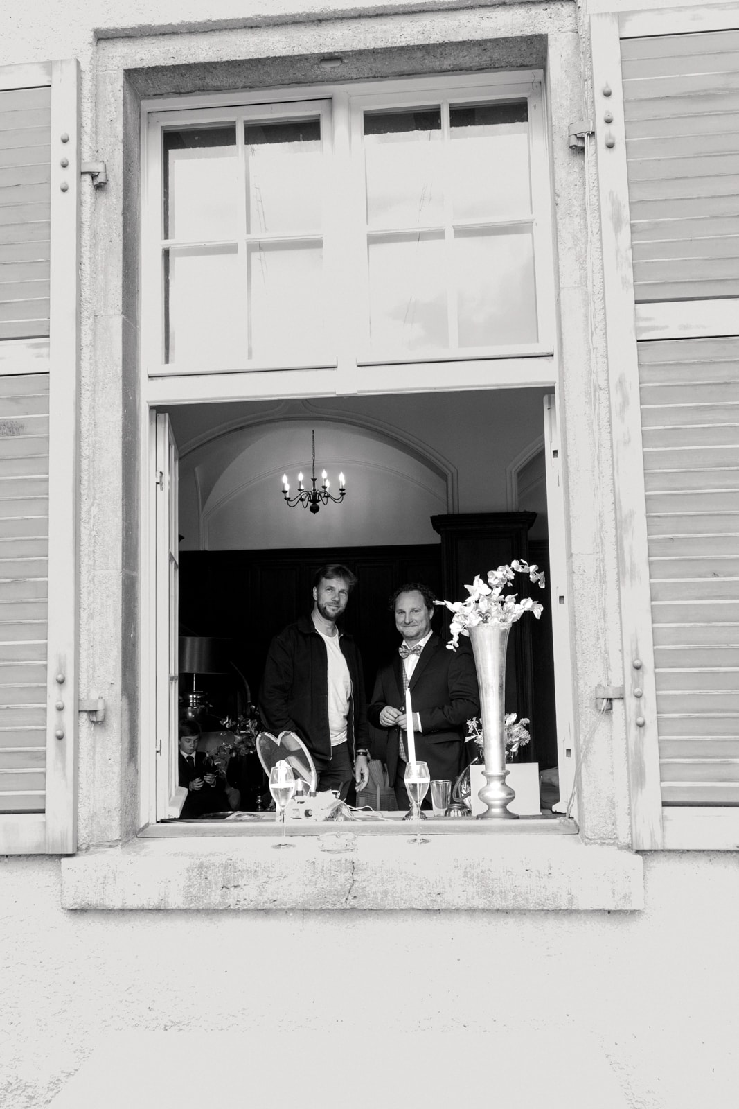 Wedding guests in formal attire standing at historic mansion window with chandelier