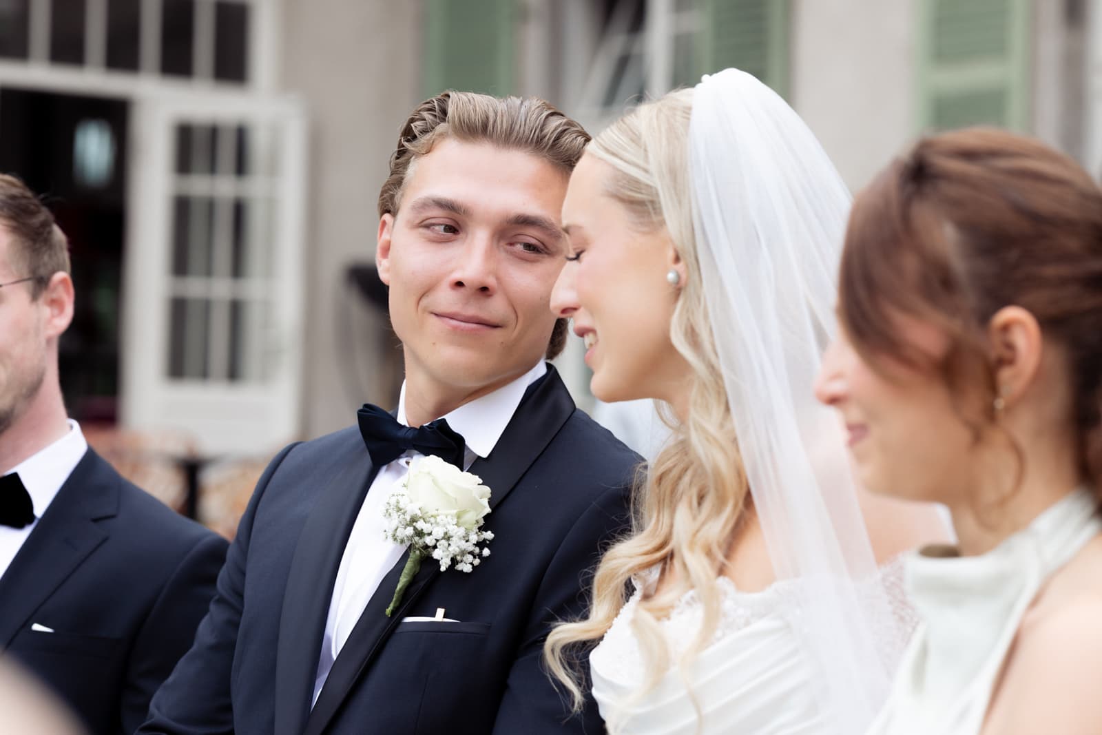 Bräutigam im schwarzen Anzug mit Fliege und weißer Ansteckblume schaut emotional zur Seite während standesamtlicher Trauung, Braut mit Schleier lächelt neben ihm
