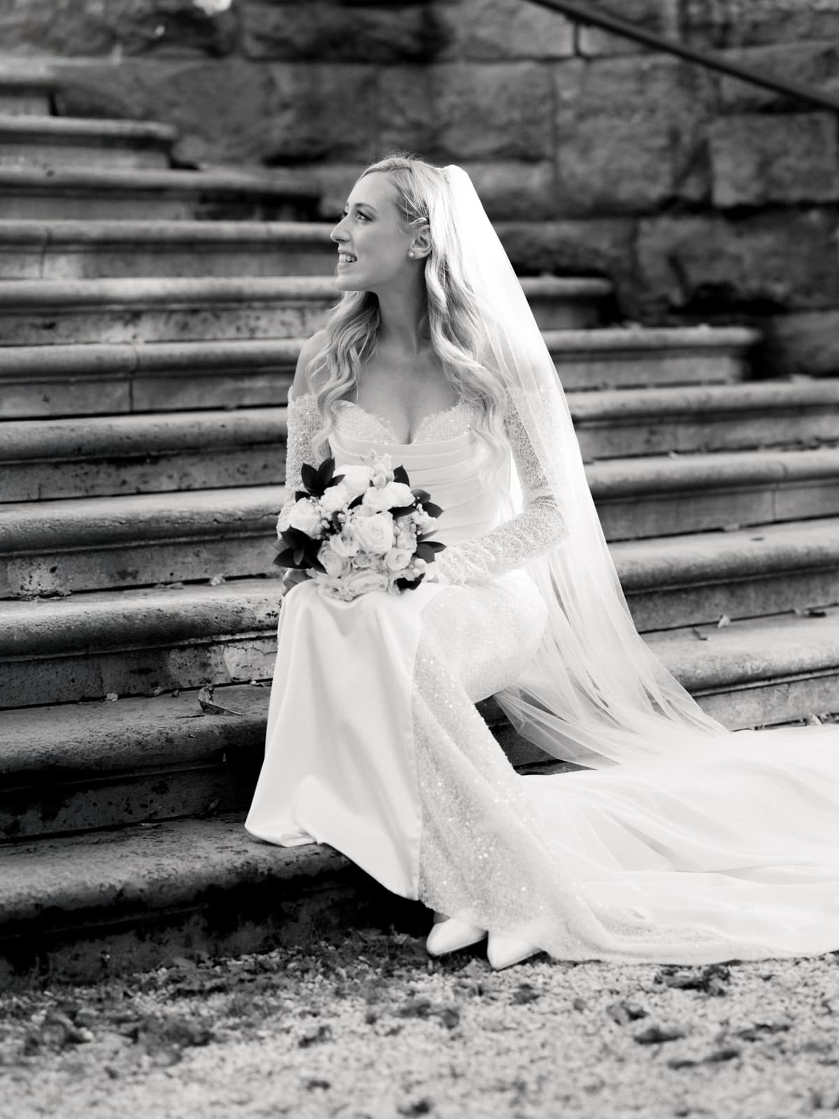 Braut in elegantem Spitzenkleid mit Schleier und Brautstrauß auf Steintreppe - Hochzeitsfotografie Schloss Kartzow