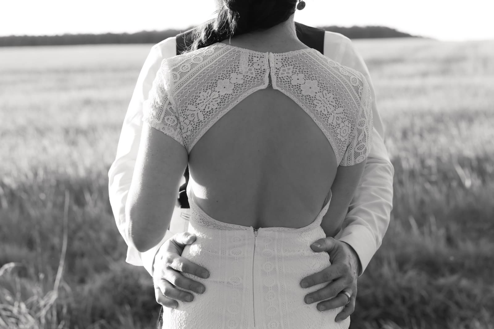 Brautpaar umarmt sich von hinten fotografiert in einem Kornfeld, Braut trägt Kleid mit Spitzendetails, Hochzeitsfotografie Brandenburg