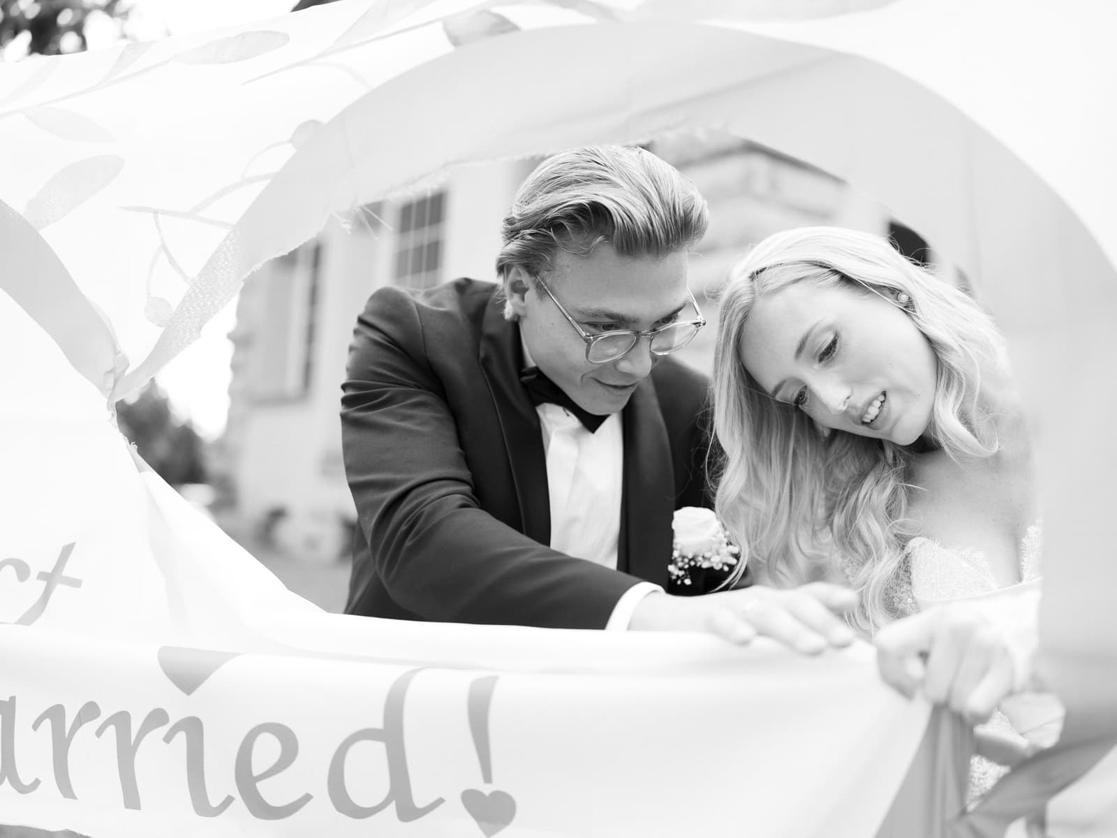 Brautpaar schaut lächelnd durch herzförmiges Just Married Schild bei Hochzeit Schloss Kartzow, schwarz-weiß