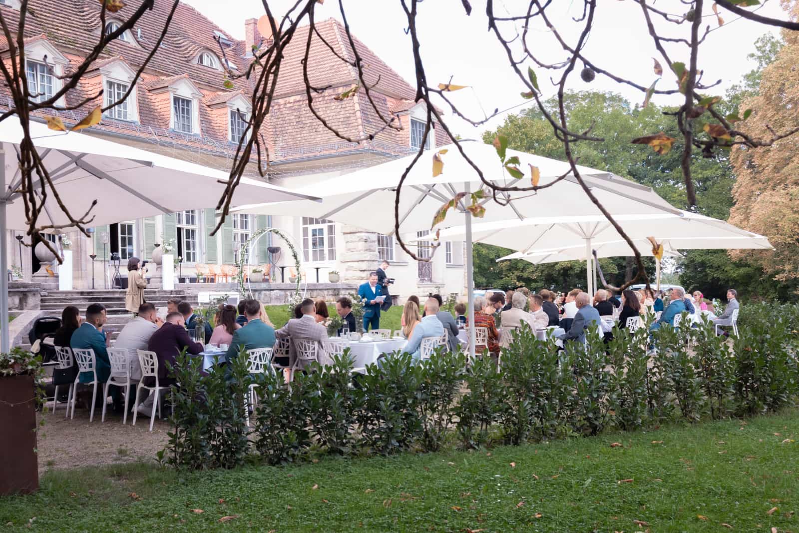 Hochzeitsgäste an Tischen unter Sonnenschirmen vor Herrenhaus durch Zweige fotografiert
