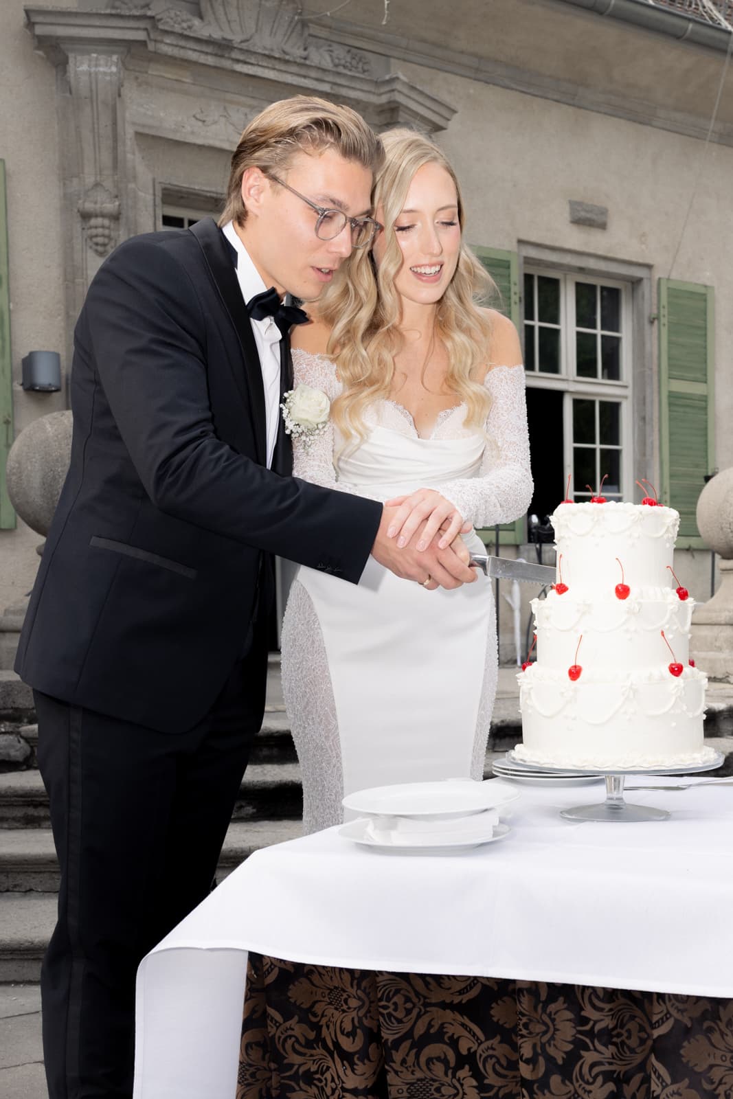 Brautpaar schneidet gemeinsam weiße Hochzeitstorte mit roten Kirschen vor historischem Gebäude an